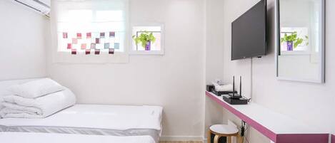 A hotel room with two beds, a desk with a computer and a chair, a flat-screen TV, and a window with a view of the city.