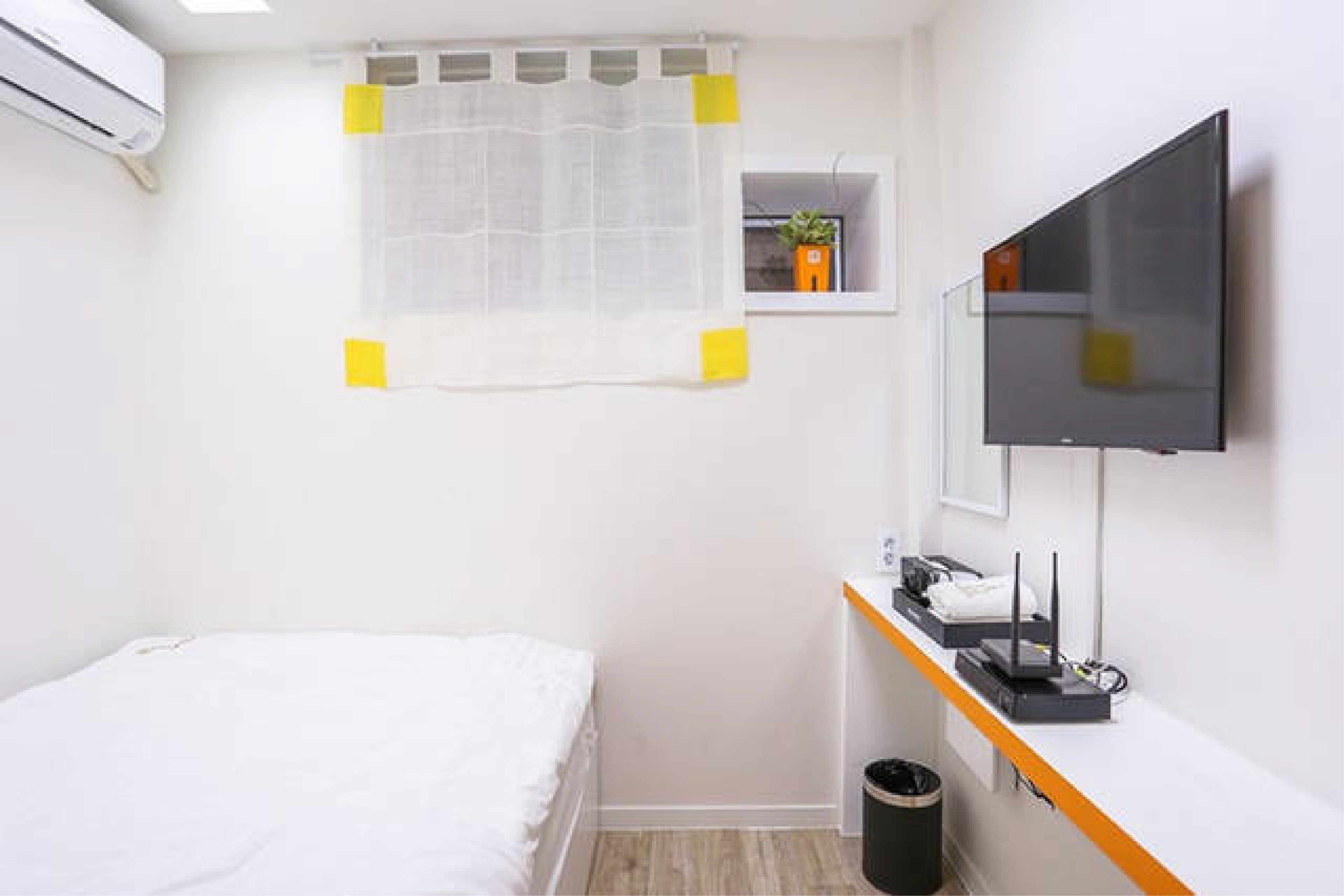 A hotel room with a bed, a small window with a sheer curtain, a TV mounted on the wall, a desk with a computer and a trash bin.
