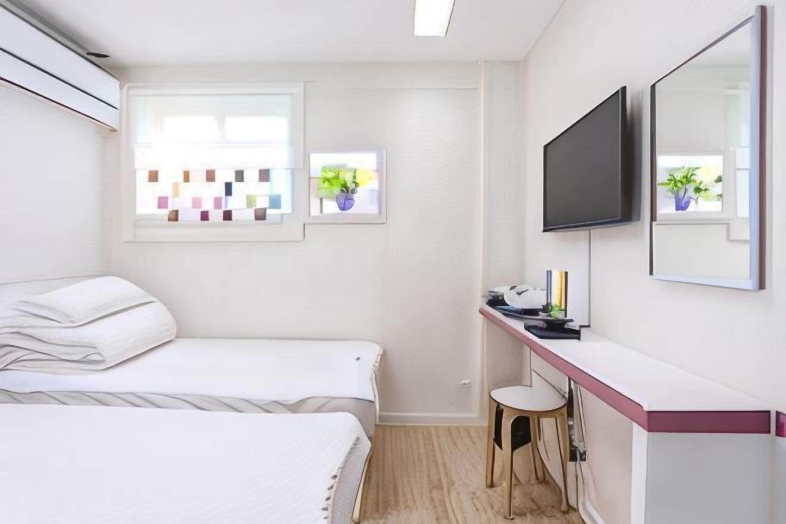 A bedroom with two beds, a desk, a chair, a TV, and a window with a view of plants.