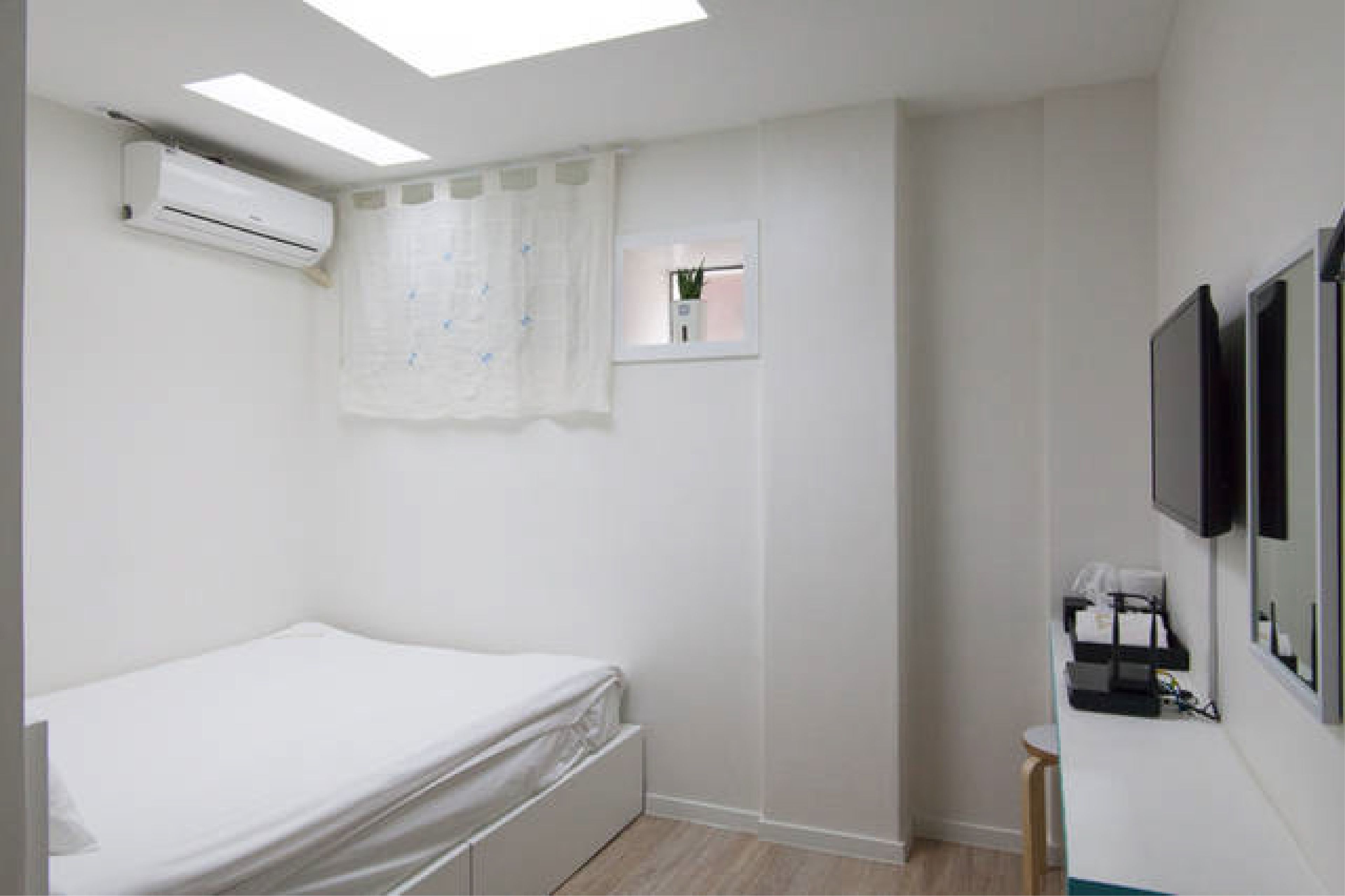 A bedroom with a bed, an air conditioning unit, a window with a curtain, a small window, and a television mounted on the wall.
