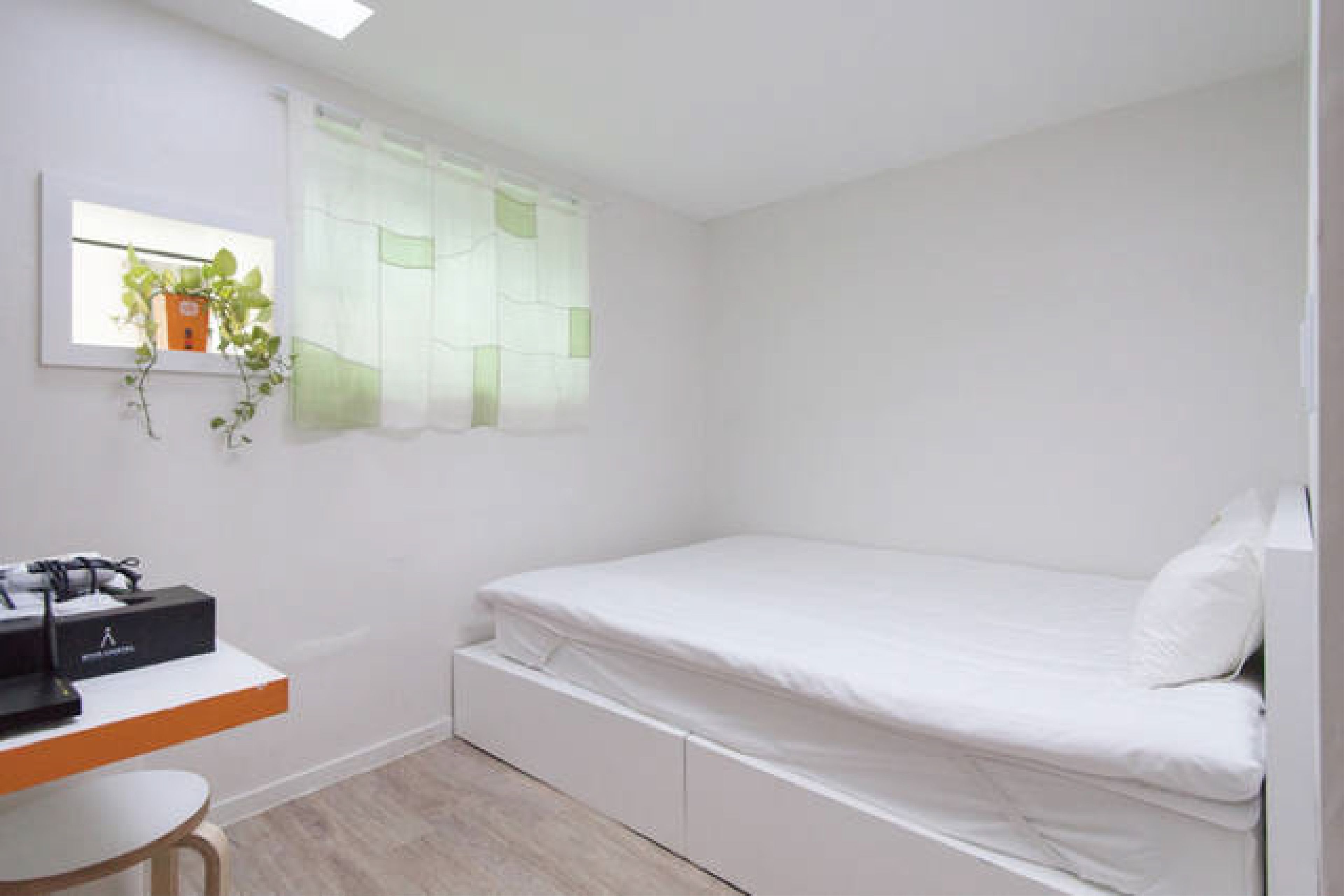 A small, clean room with a bed, a desk, and a window with a plant.