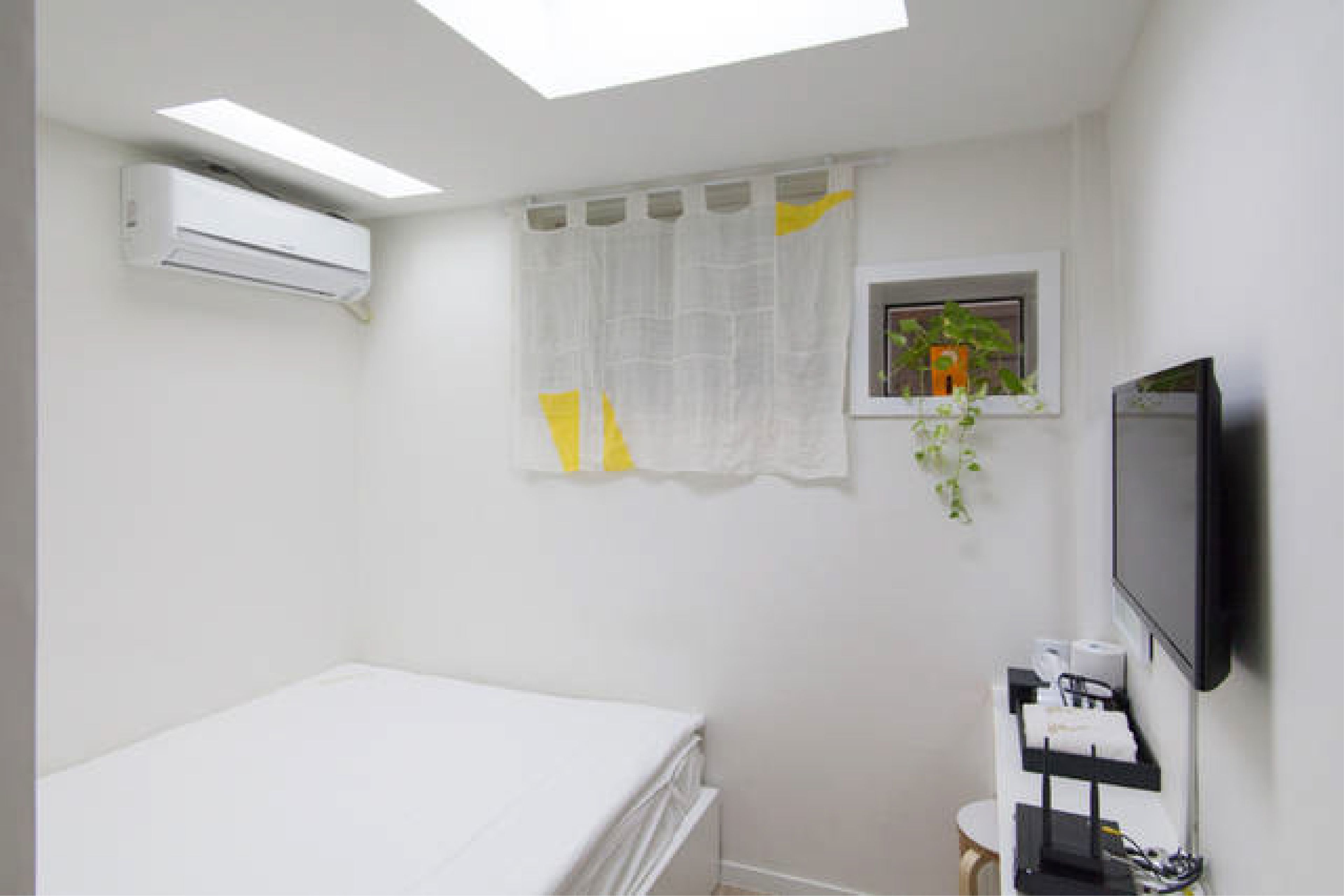 Un dormitorio con cama, aire acondicionado, ventana con cortina, planta, televisor y mesita de noche con artículos de aseo.
