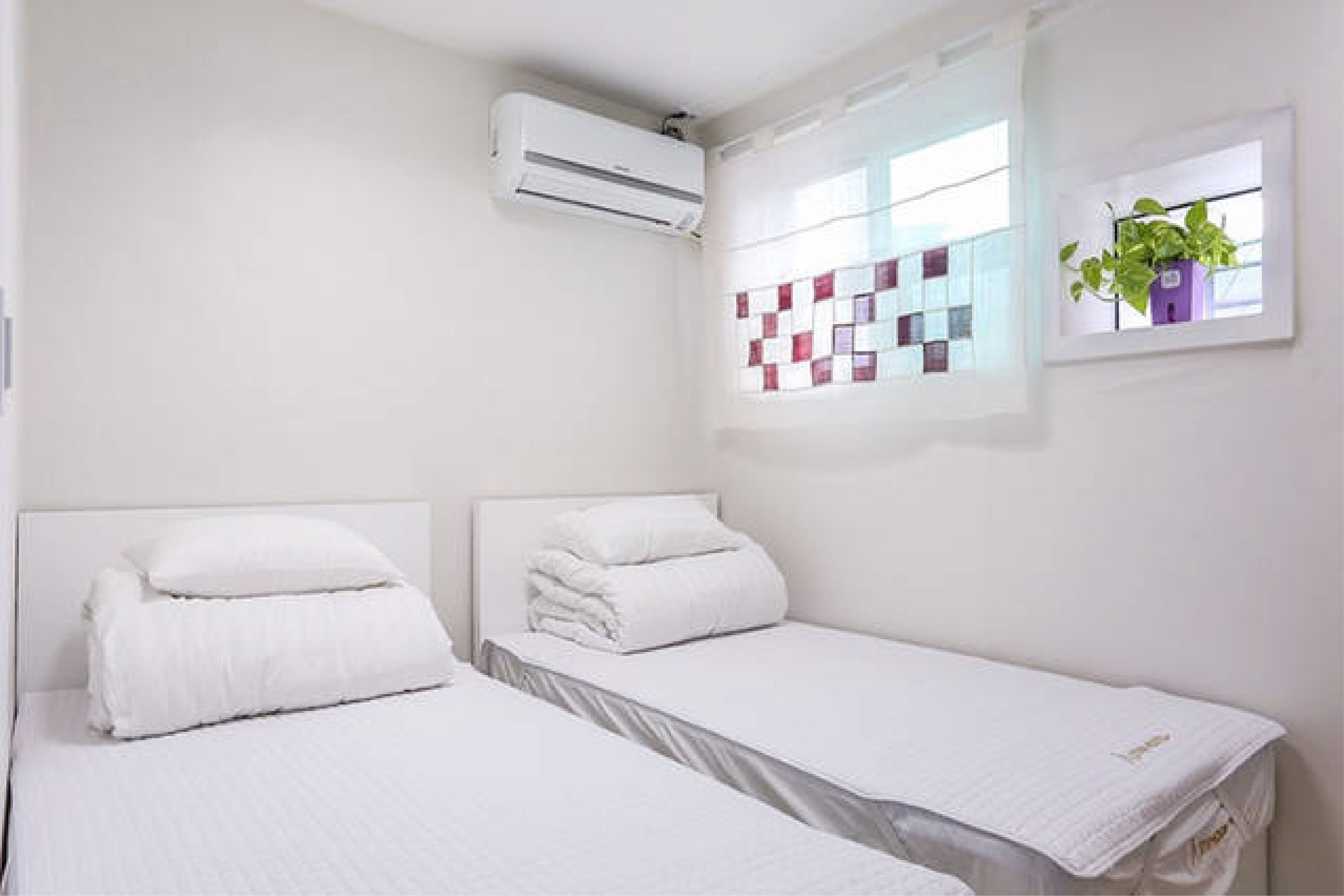 A hotel room with two beds, an air conditioning unit, a window with a view of the city, and a plant on the windowsill.