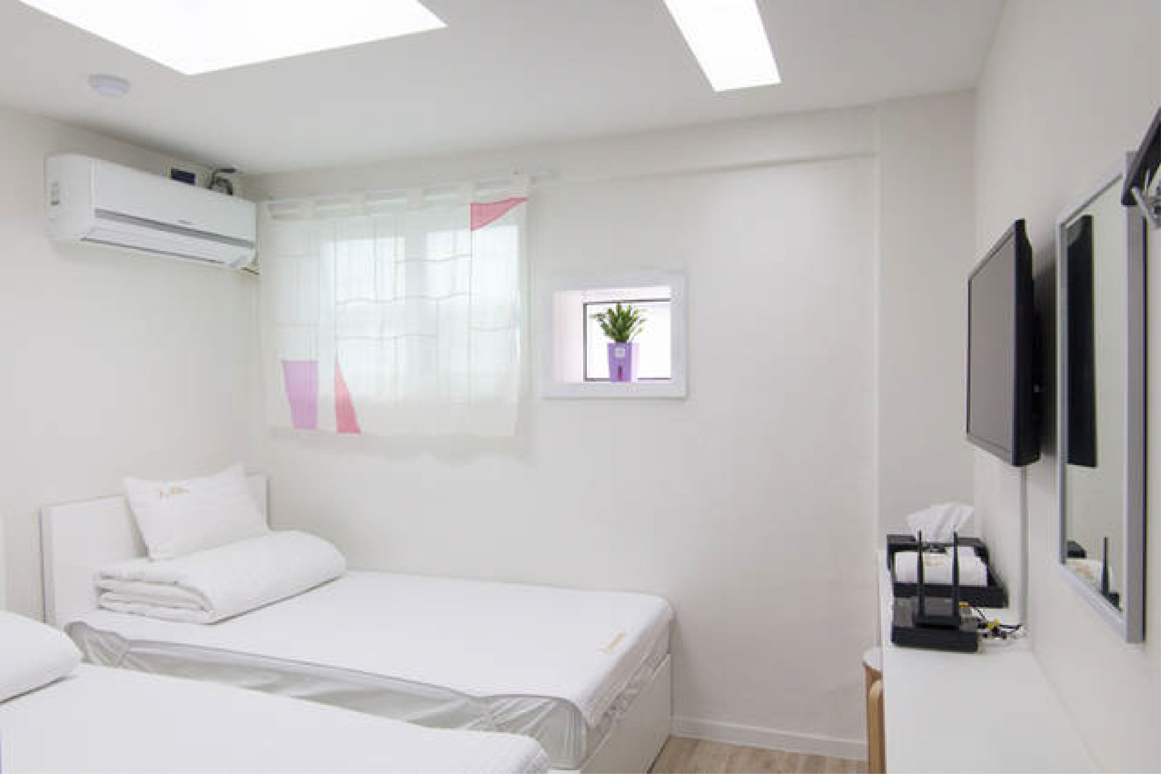 A hotel room with two beds, an air conditioning unit, a small window with a plant, and a television mounted on the wall.