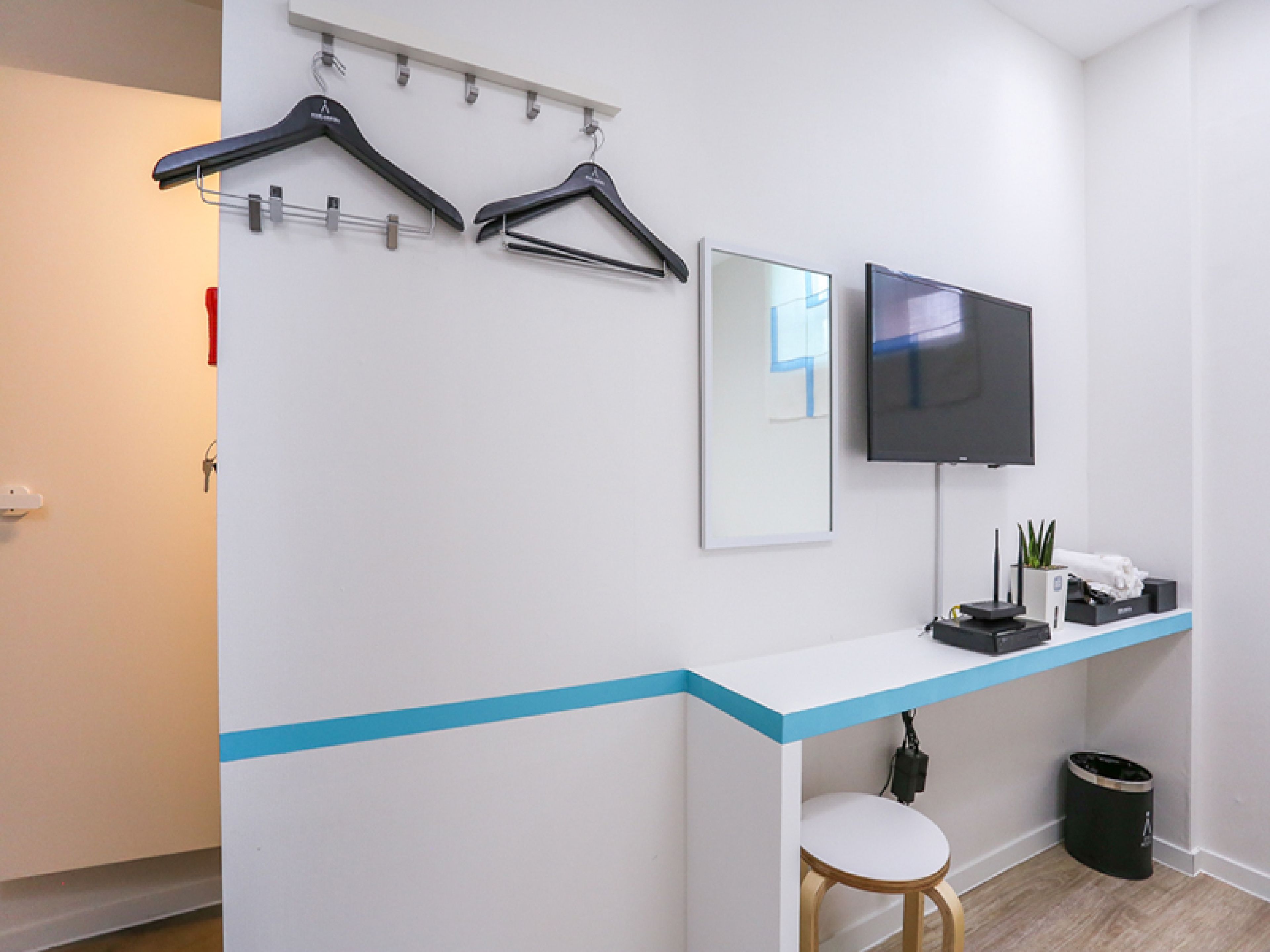 A hotel room with a white wall, a blue accent strip, a mounted TV, a small table with a chair, and a trash can.