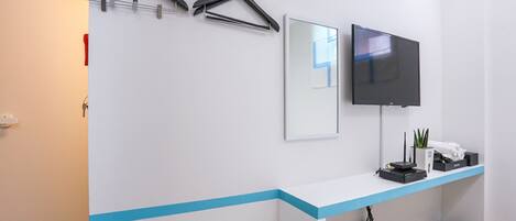 A hotel room with a white wall, a blue accent strip, a mounted TV, a small table with a chair, and a trash can.