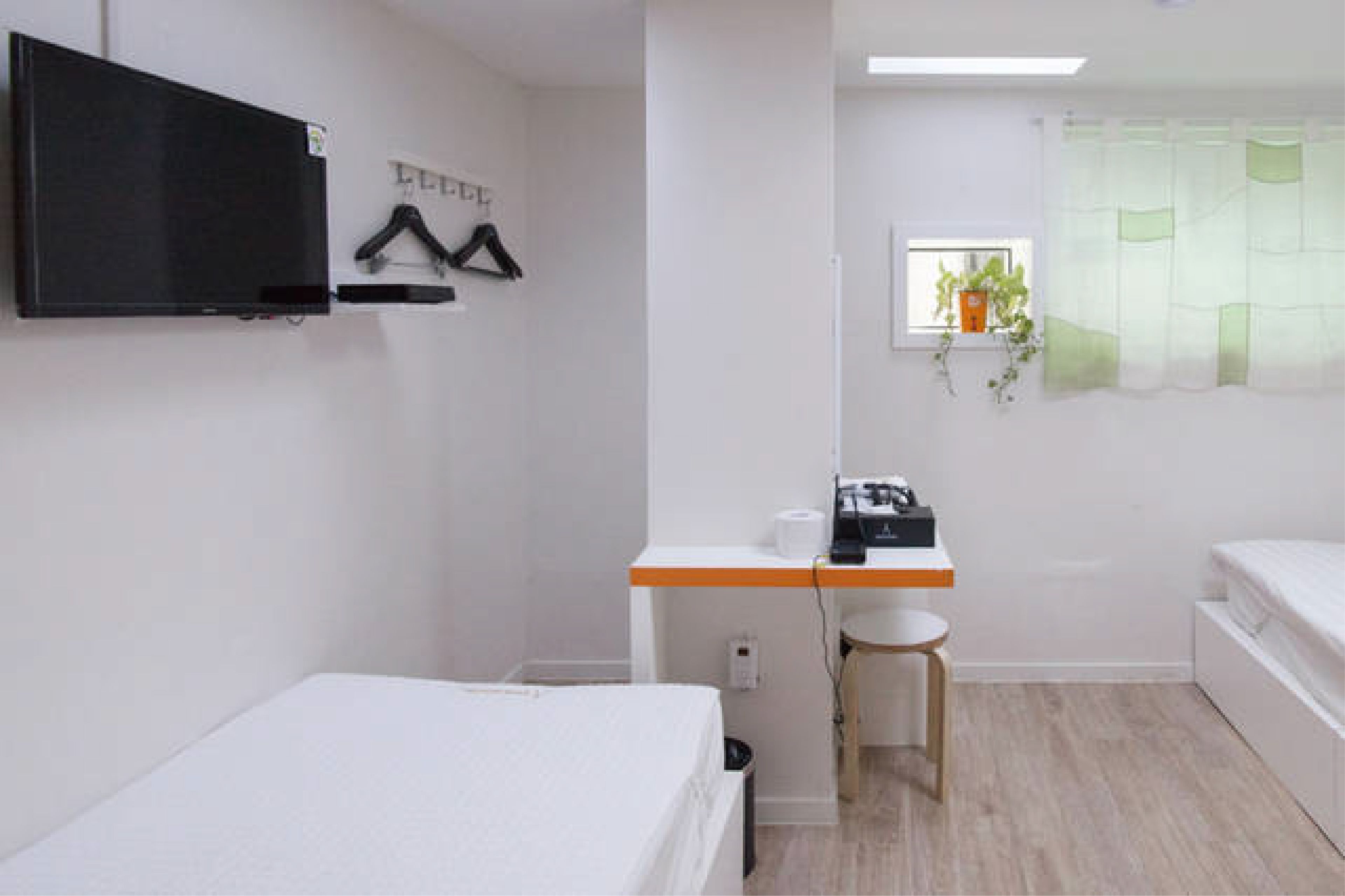 A small, clean room with a bed, a desk, a chair, a TV, and a window with a plant.