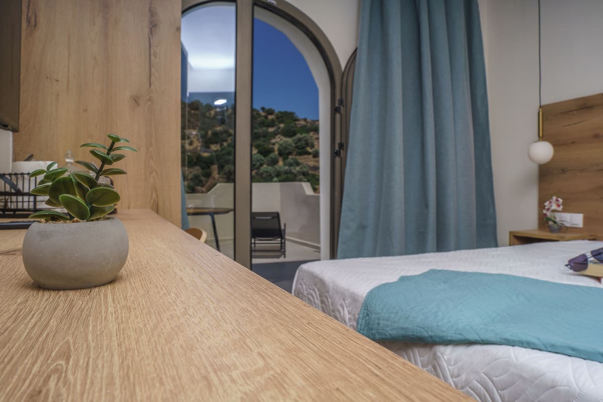 Un dormitorio con una cama, una mesa de madera, una planta en maceta, una mesita de noche y vistas al exterior.