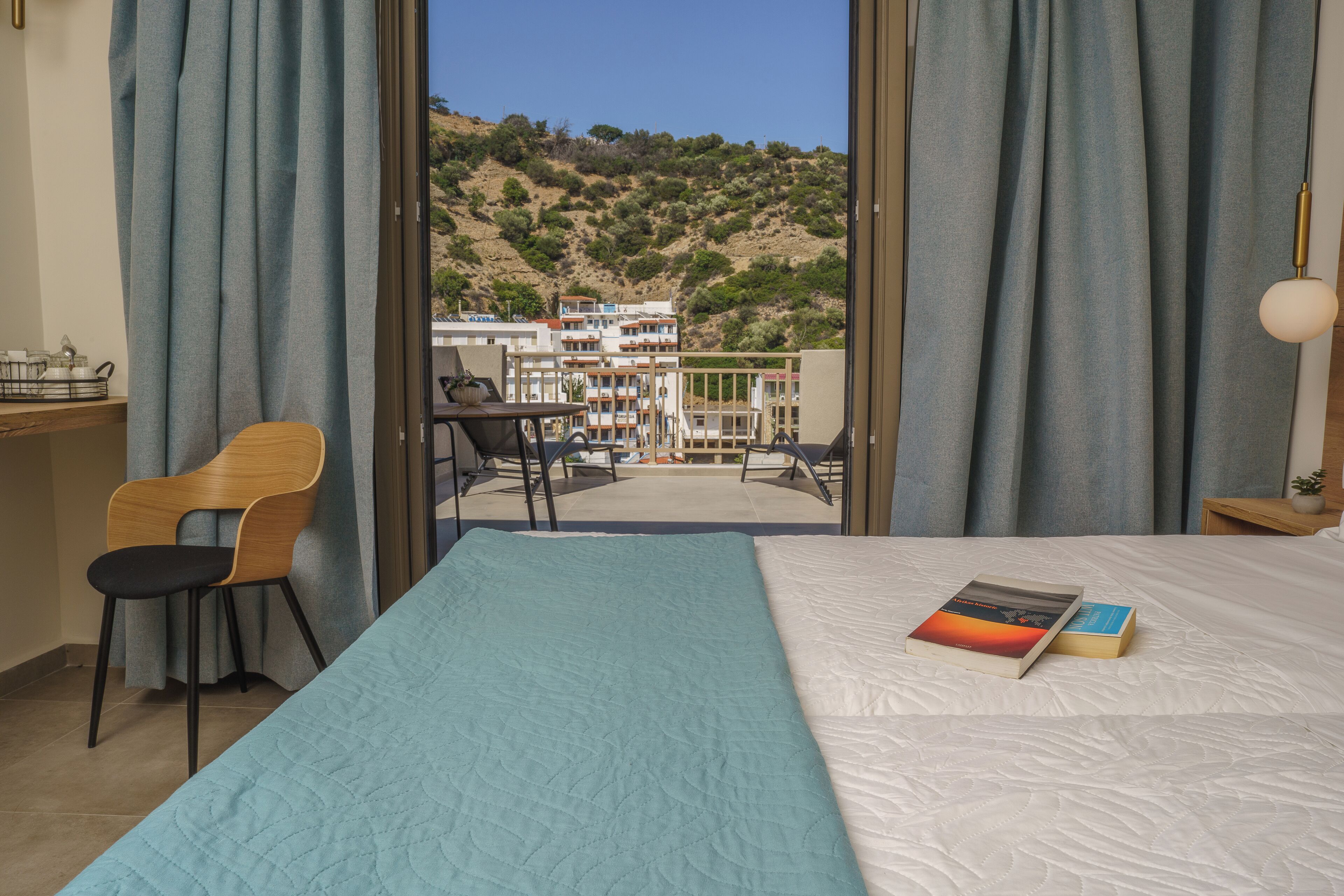 A hotel room with a bed, a chair, a desk, and a view of a building and hills.
