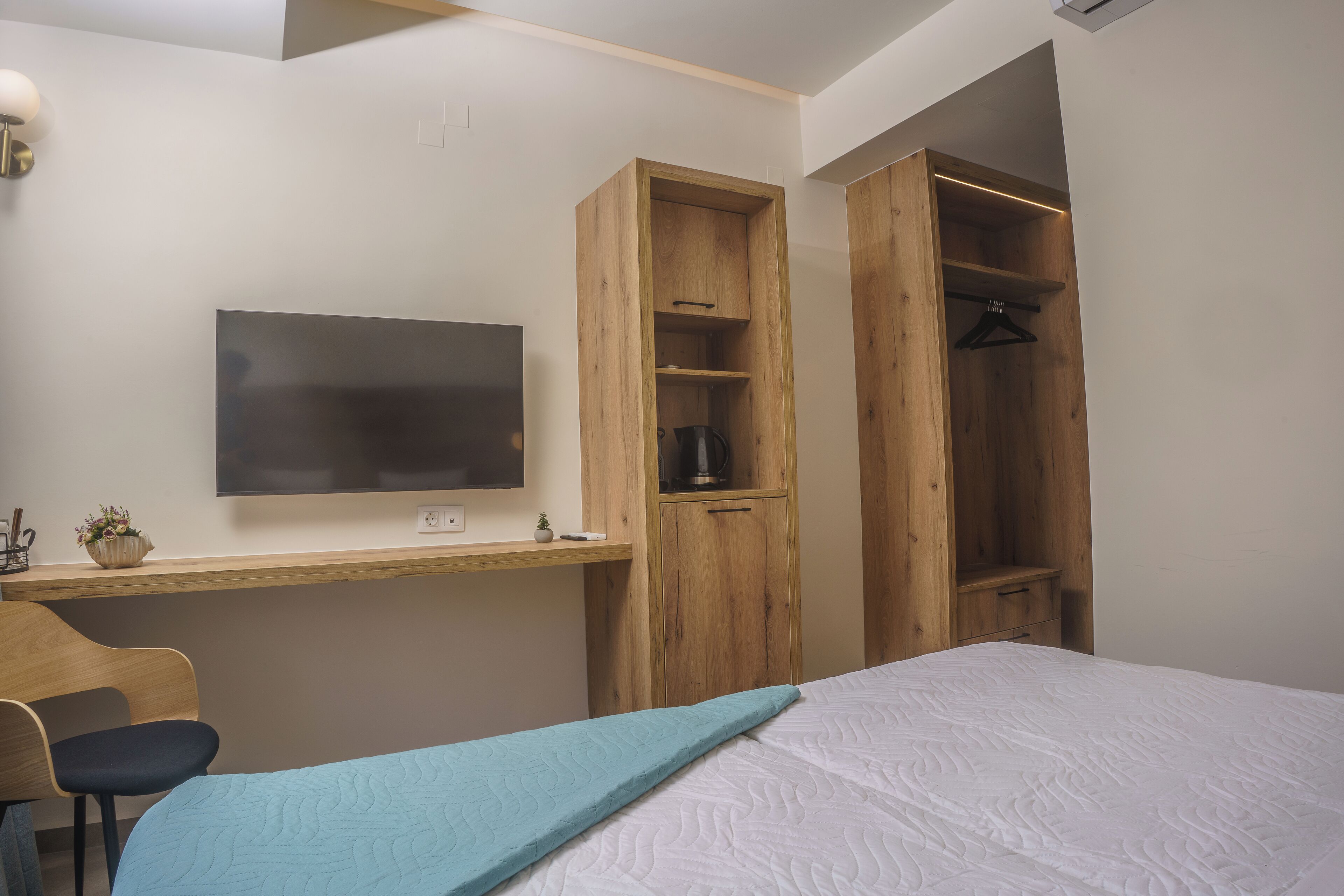 A bedroom with a bed, a desk with a TV, a wooden wardrobe, and a hanging closet.