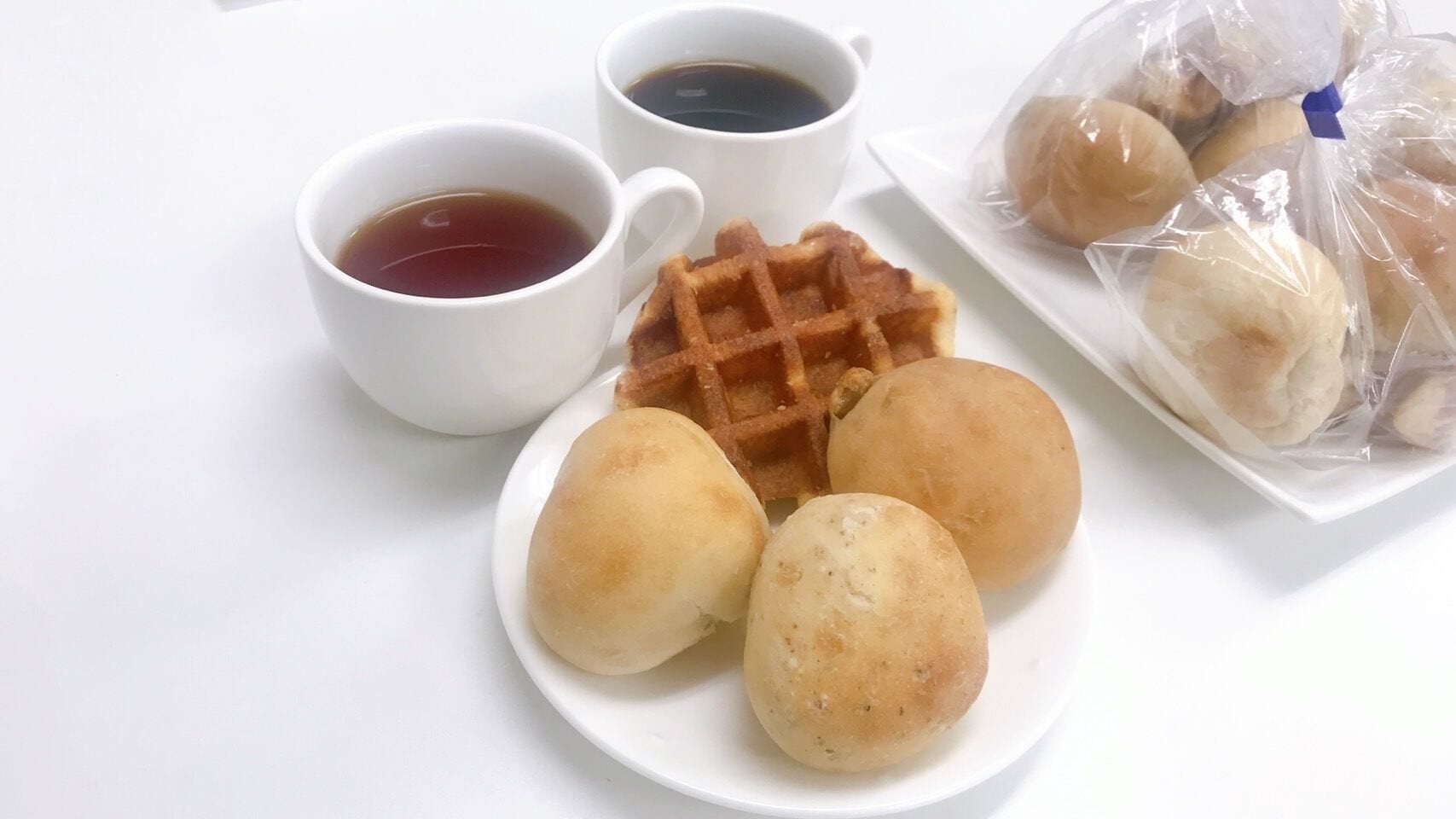 A plate with three round buns, a waffle, and two cups of tea on a white table.