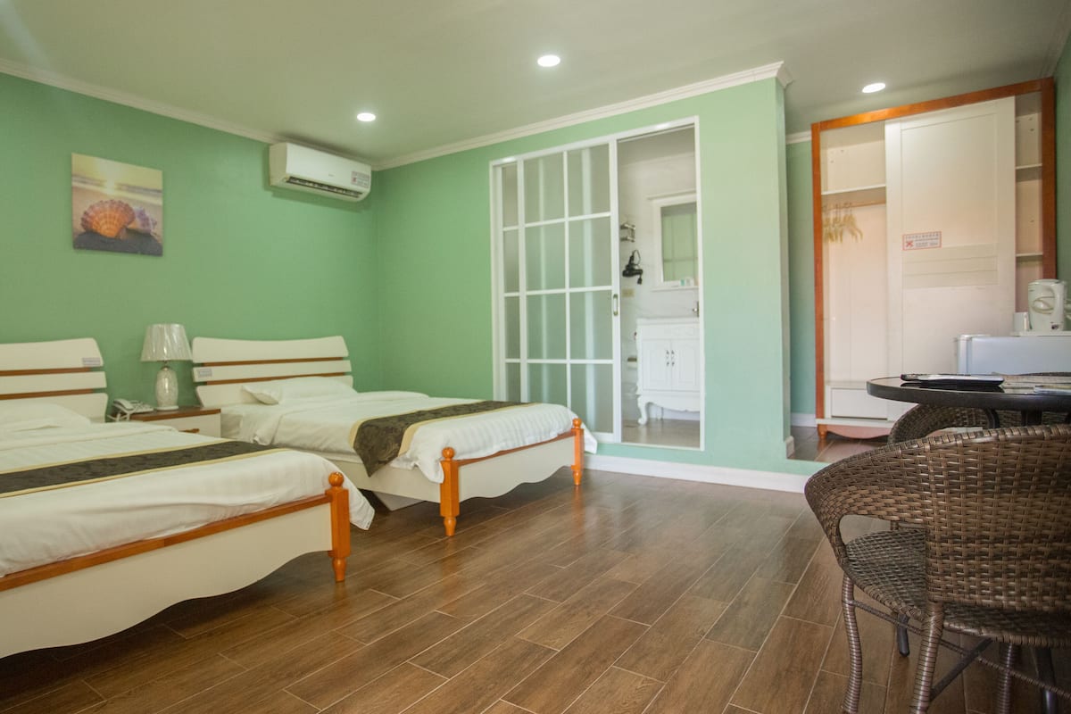 A hotel room with two beds, a wicker chair, a desk, and a bathroom visible through a glass door.