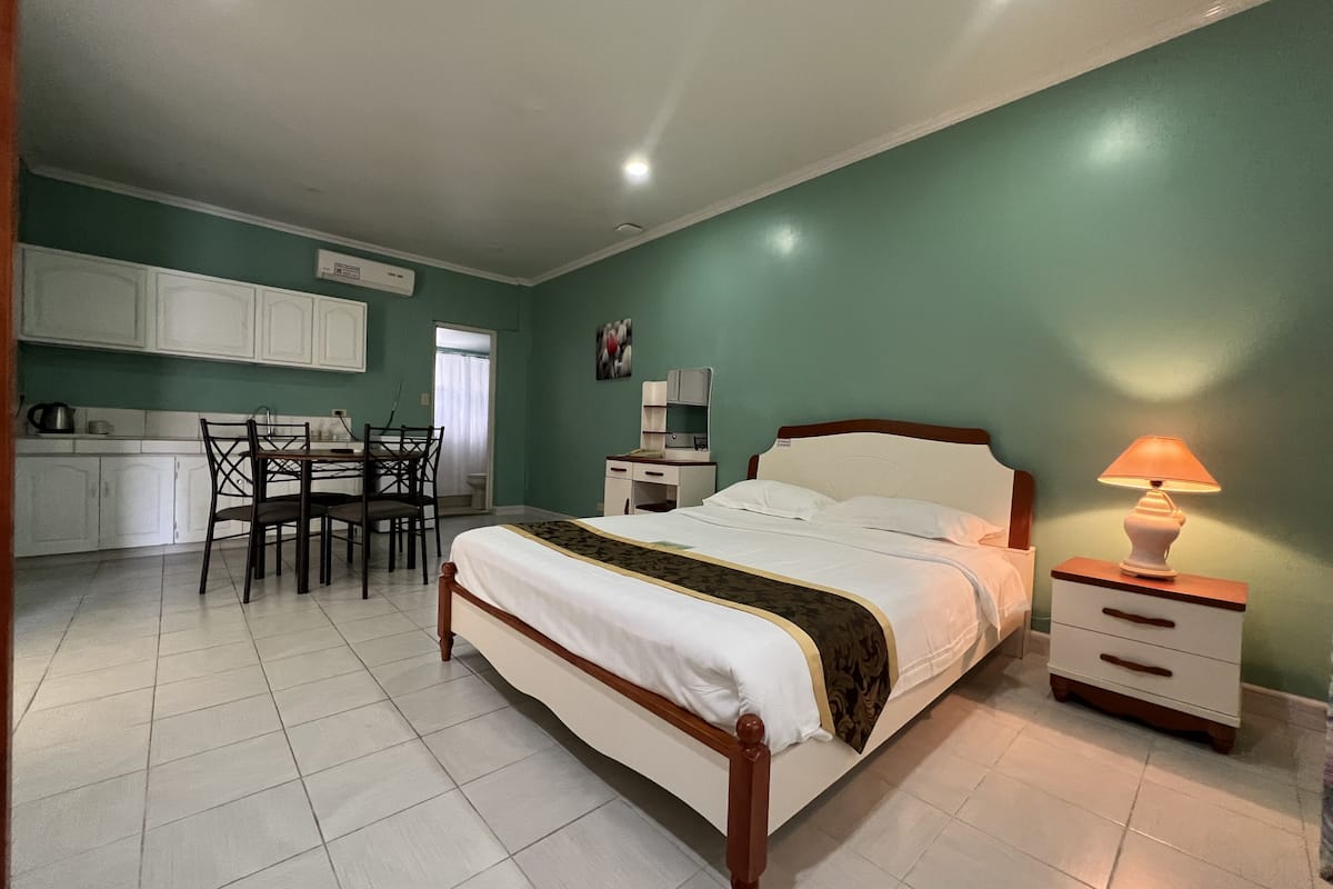 A bedroom with a bed, bedside table, and lamp. There is a dining area with a table and chairs. The room has green walls and white flooring.