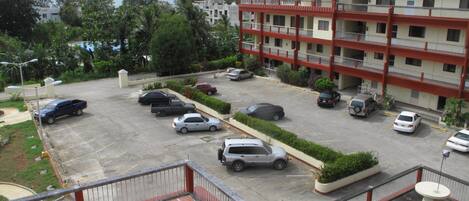 A parking lot with several cars, a multi-story building with balconies, and surrounding greenery.