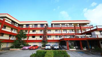 A multi-story building with a red and beige color scheme, featuring balconies and a parking area with several cars.