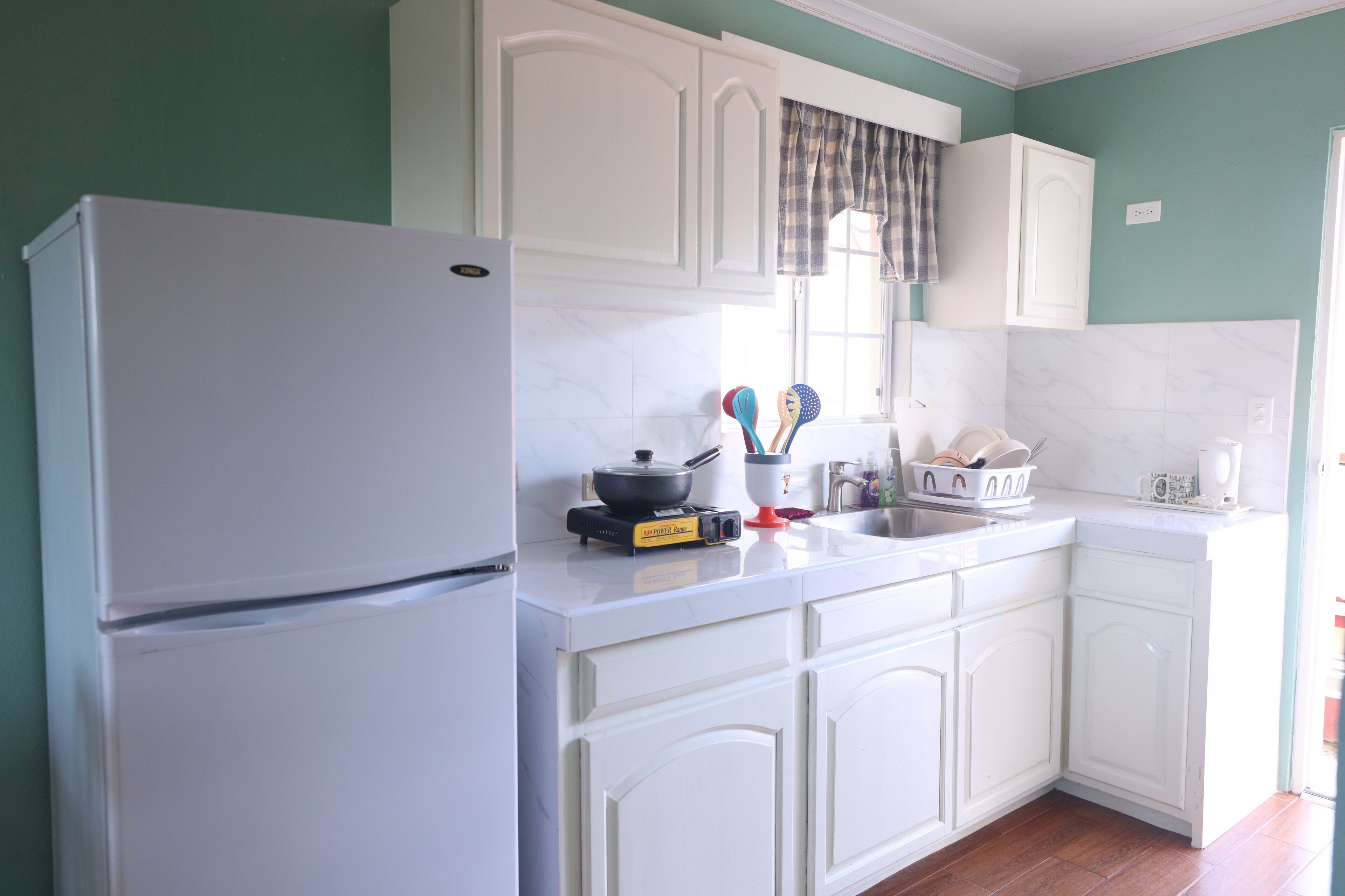 Una cocina con armarios blancos, un refrigerador blanco y una ventana con cortina escocesa.