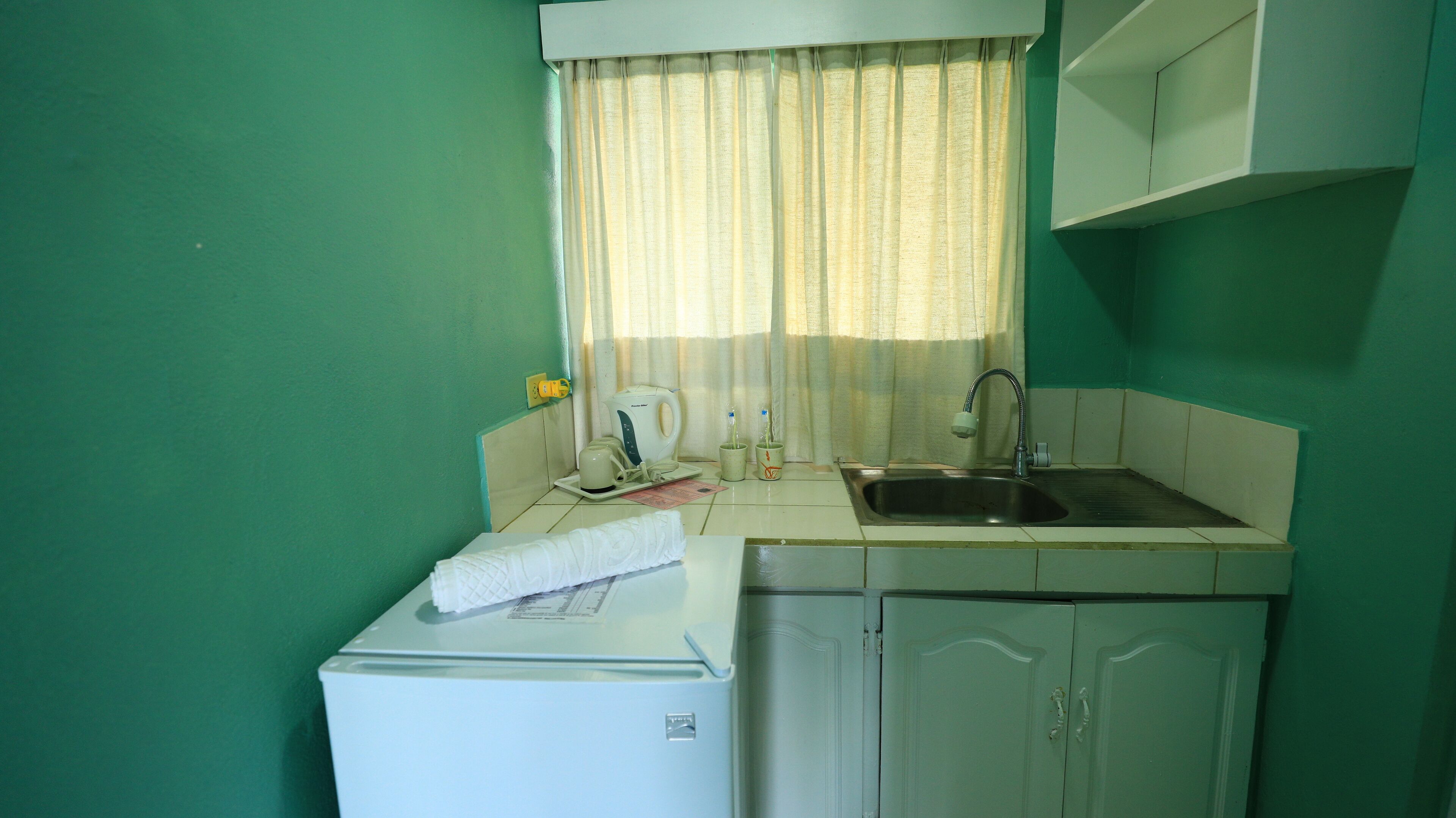A compact kitchen with a refrigerator, a sink, and a window with curtains.