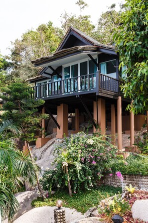 A two-story house with a balcony, surrounded by tropical vegetation and rocks.