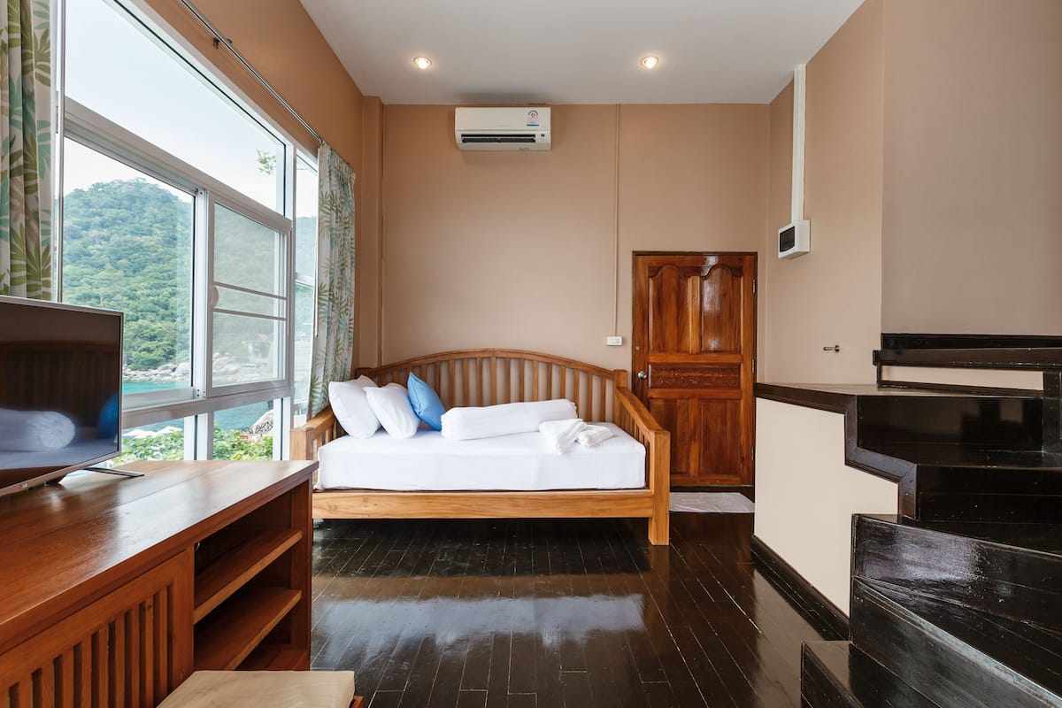 Ein Schlafzimmer mit einem hölzernen Bett, einem Fernseher auf einem Holzschrank, einem Kleiderschrank und einem Fenster mit Blick auf Grünflächen.