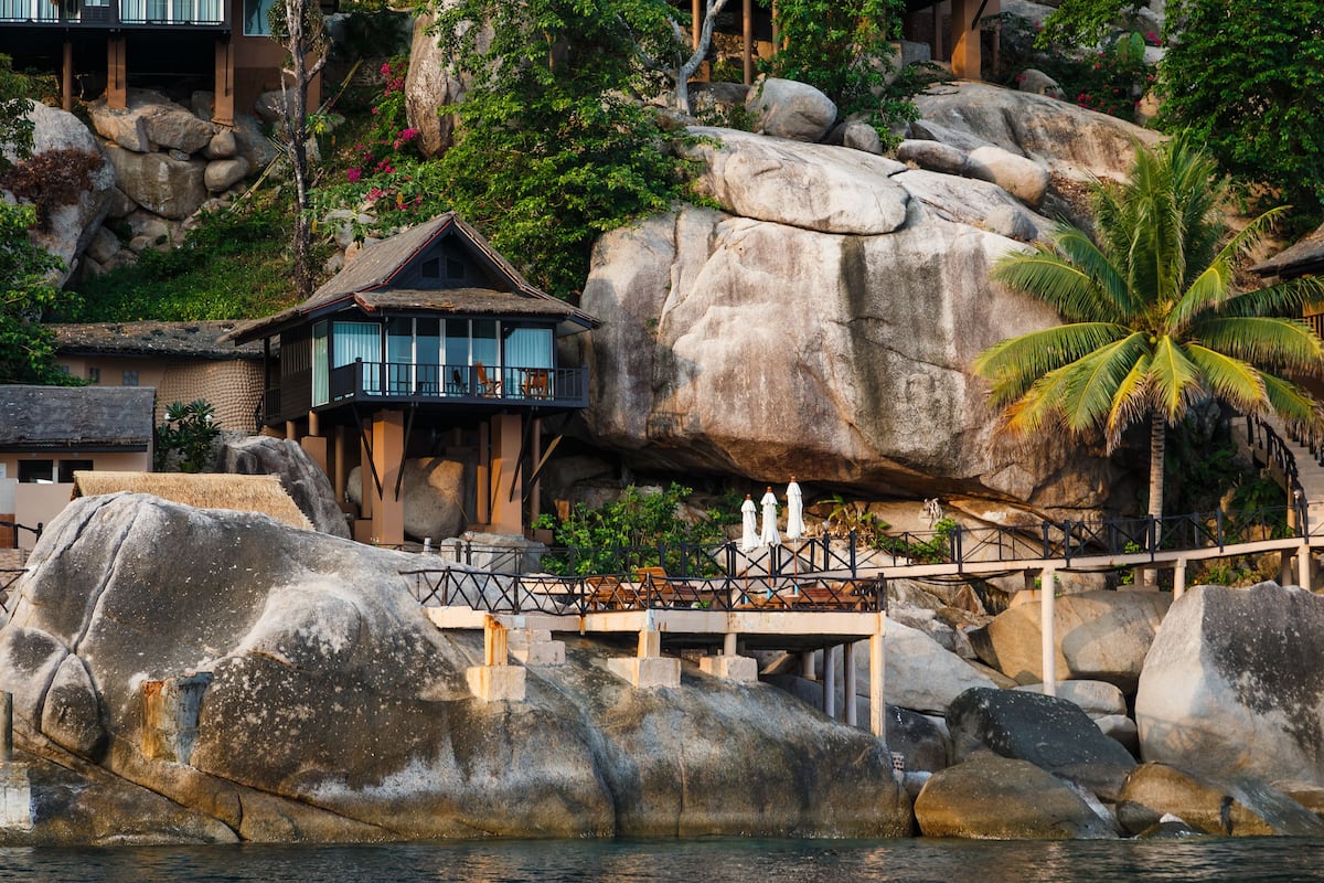 Ein Resort mit einem Gebäude mit einem Reetdach, einer Brücke und großen Felsen.