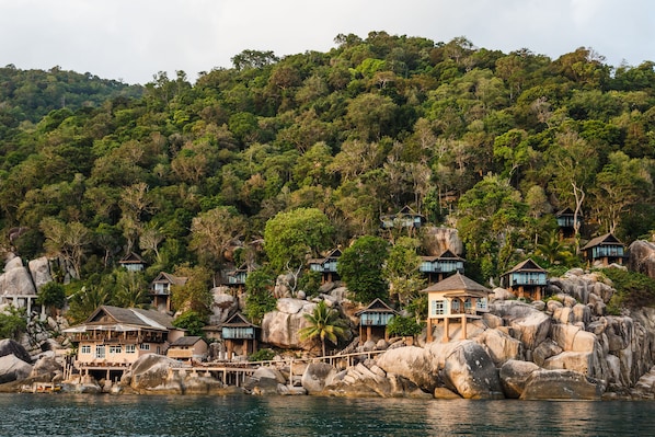 A coastal resort with multiple bungalow-style accommodations built on large rocks, overlooking a body of water and dense forest.