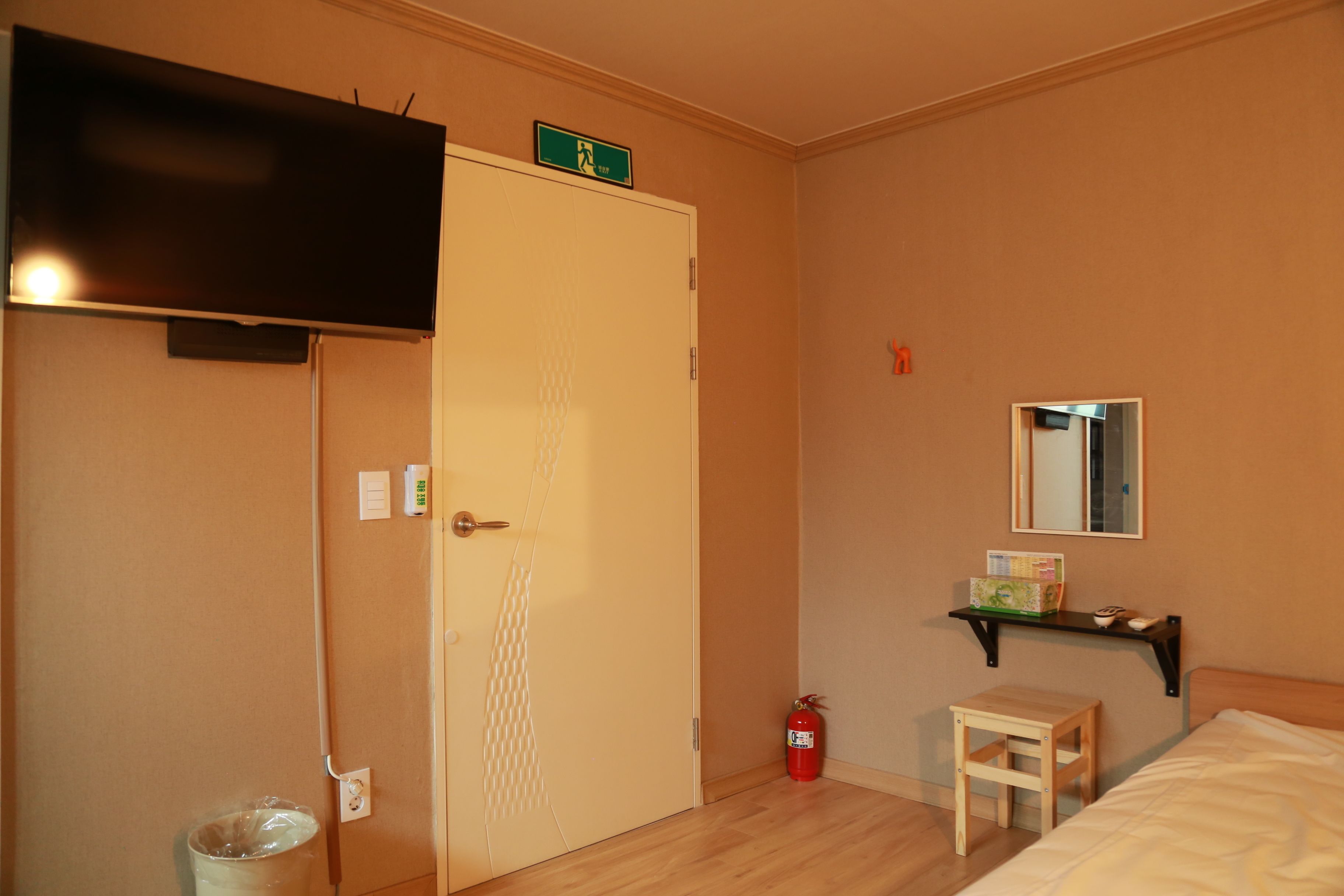Habitación de hotel con cama, una mesita, estante con objetos, extintor, puerta con letrero y televisor montado en la pared.