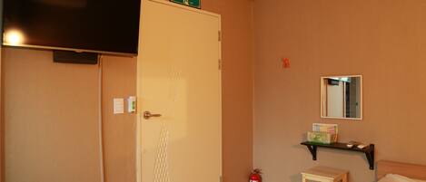 Habitación de hotel con cama, una mesita, estante con objetos, extintor, puerta con letrero y televisor montado en la pared.
