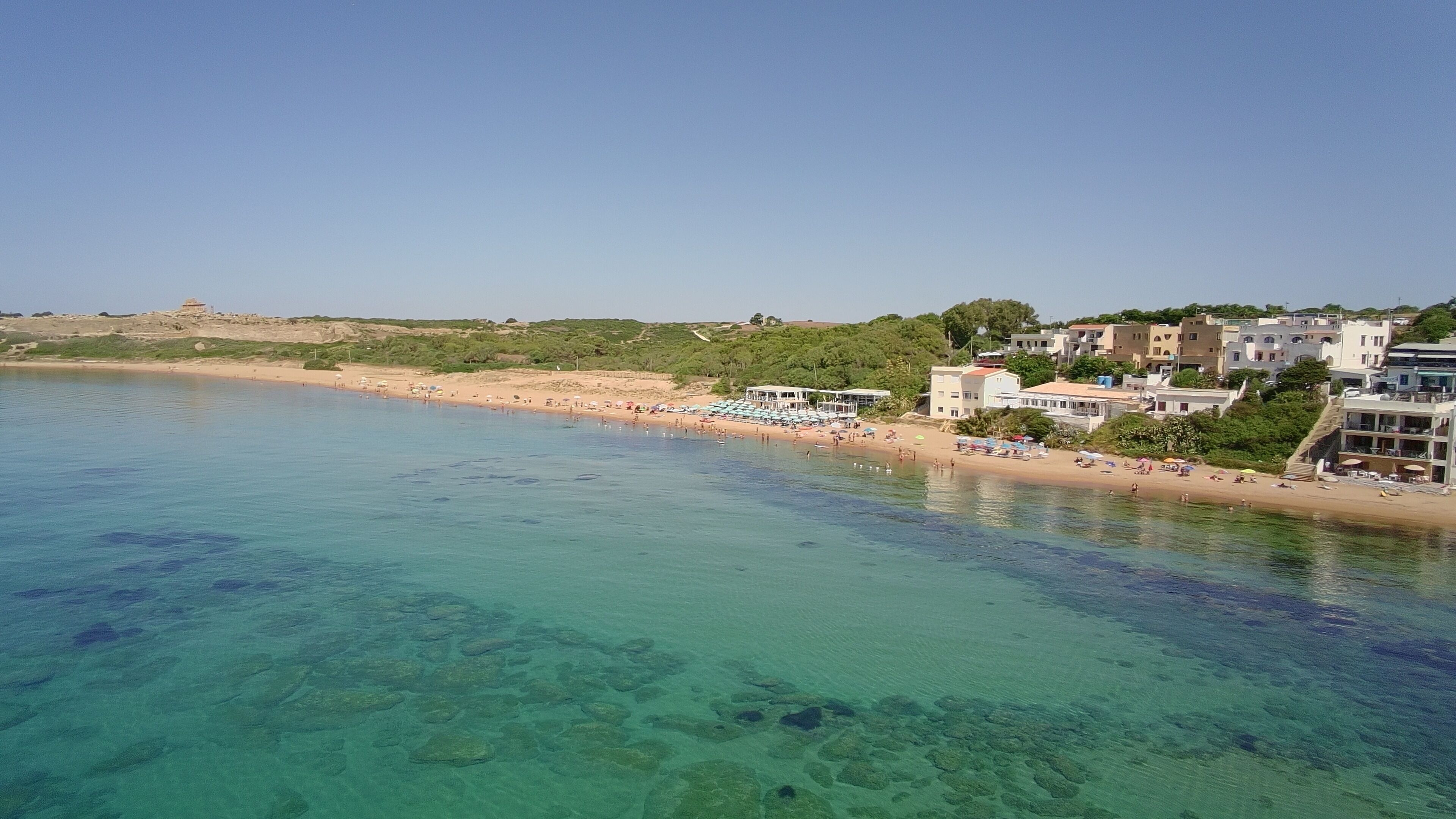 Uma área costeira com águas turquesa cristalinas, uma praia de areia e edifícios com varandas viradas para o mar.
