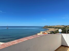 Vista al mar desde un balcón, cielo despejado, playa de arena y una colina rocosa distante con una torre.