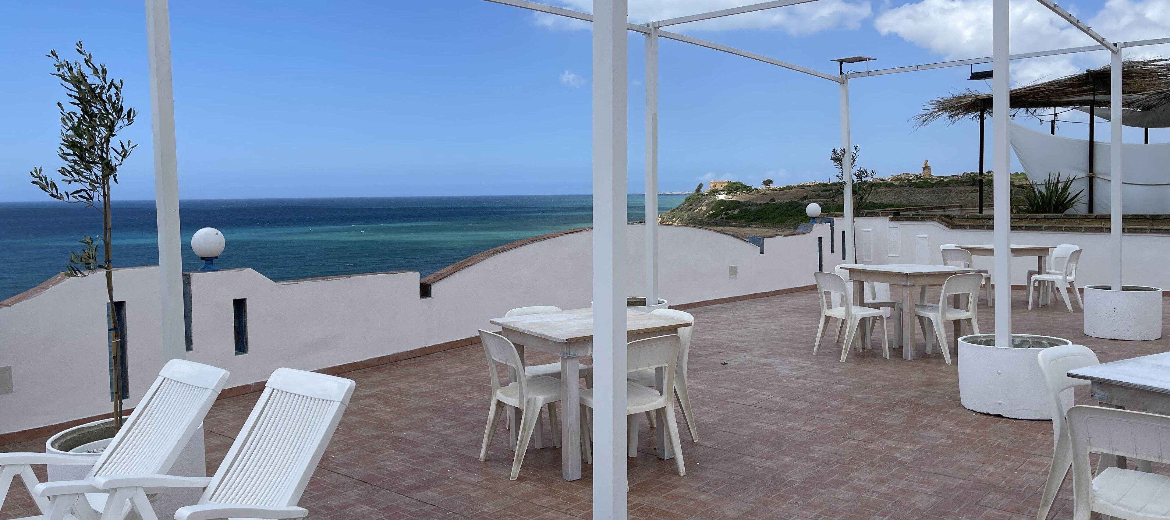 A covered outdoor seating area with white plastic chairs and tables, overlooking a coastal view.