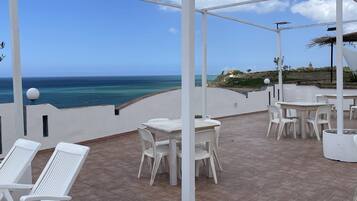 A covered outdoor seating area with white plastic chairs and tables, overlooking a coastal view.