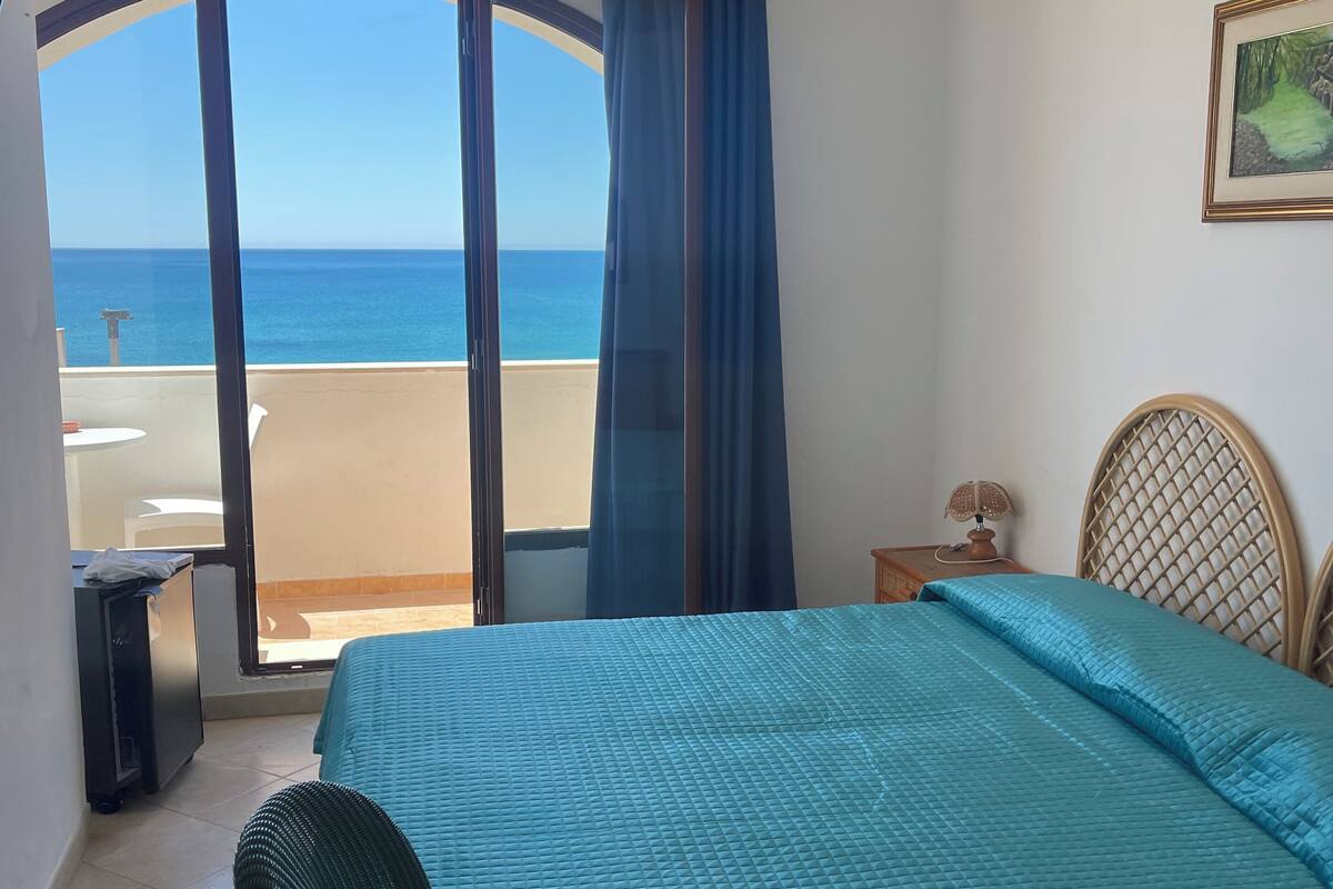 Une chambre à coucher avec un lit, une chaise, une petite table avec une lampe, un tableau au mur et une vue sur la mer par la fenêtre.