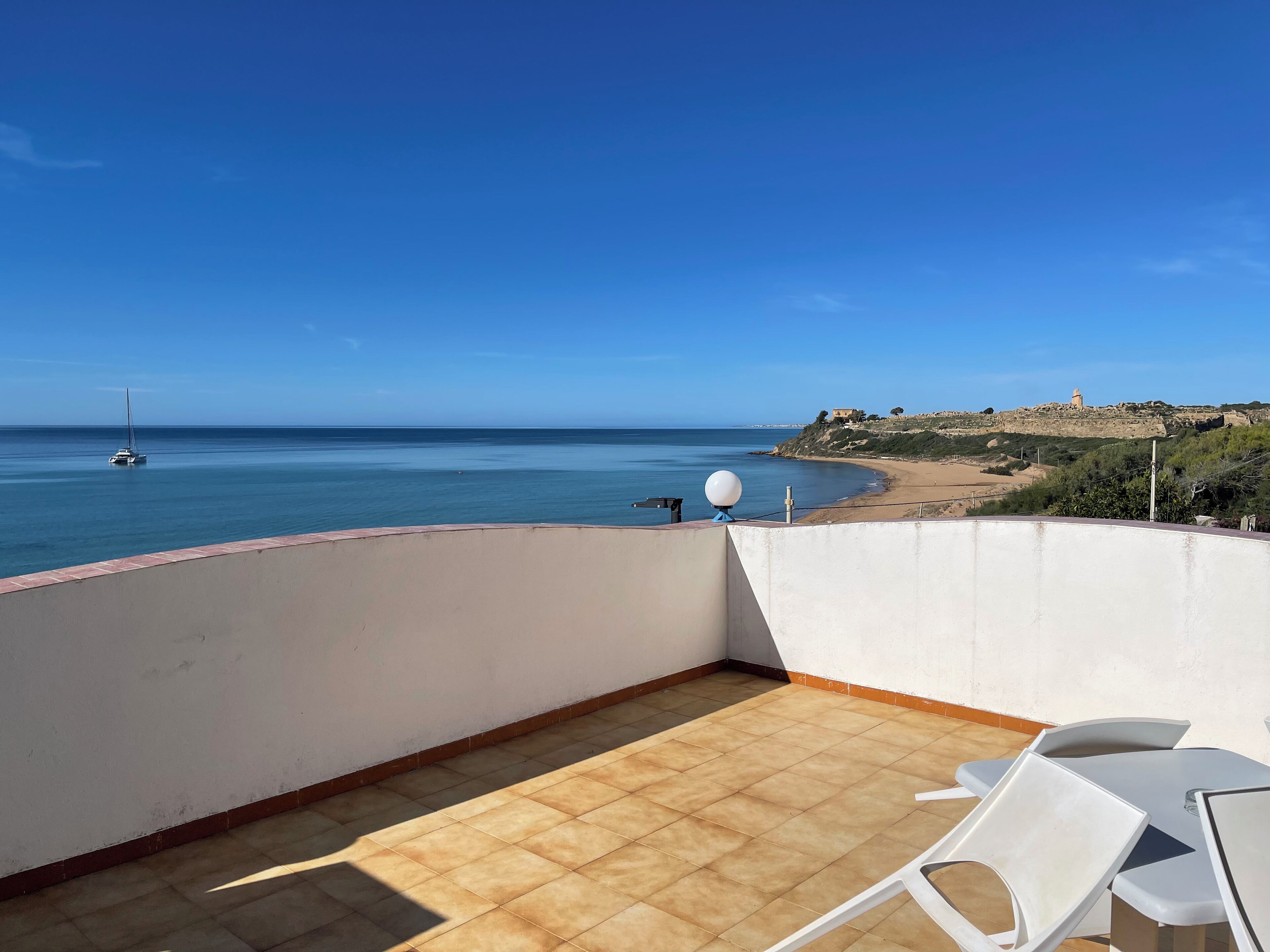 A balcony with a view of the sea, a sandy beach, and a rocky hillside with buildings.