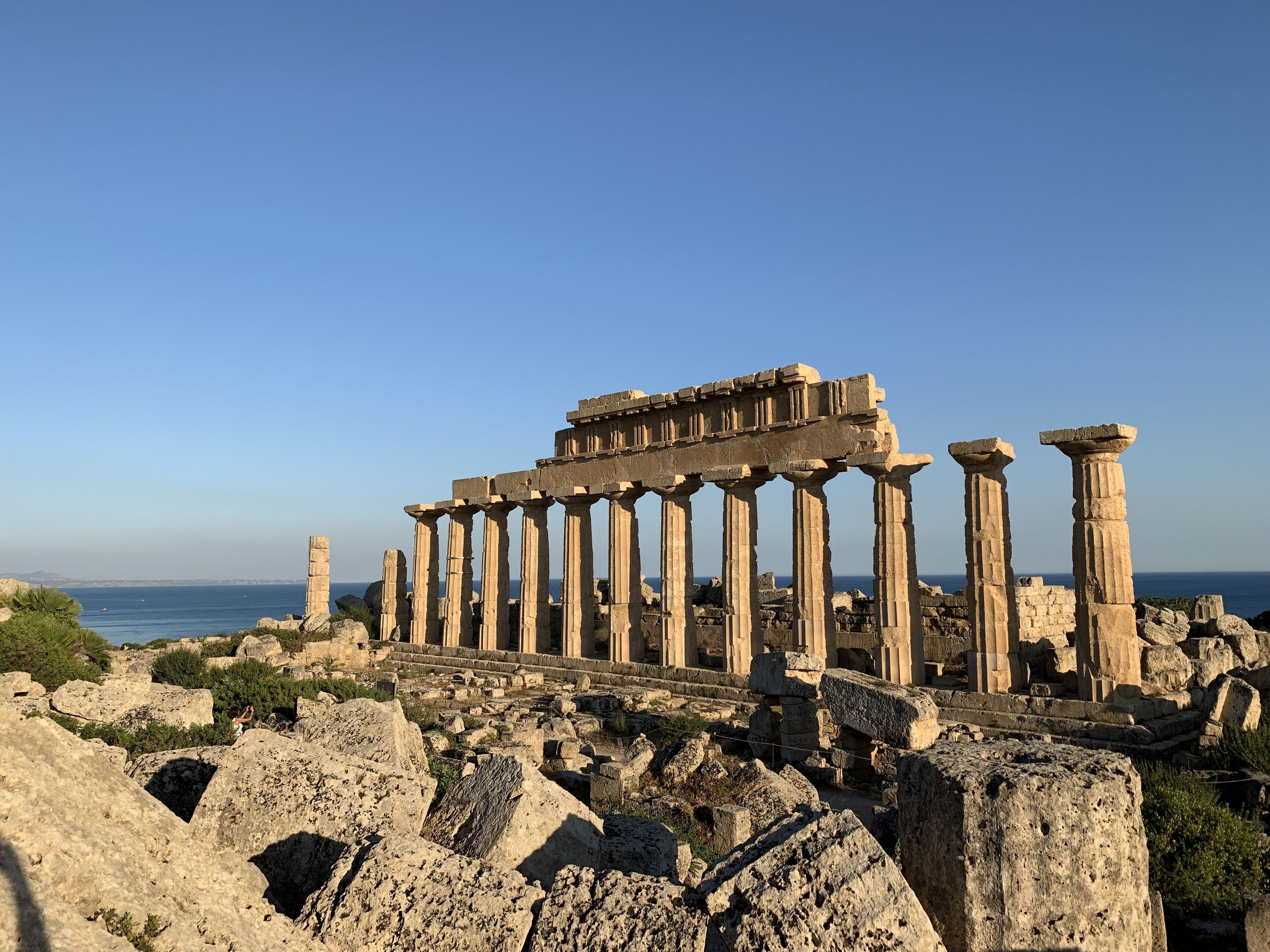 Ancient ruins with columns and a stone structure, surrounded by rocky terrain and vegetation, overlooking a body of water.
