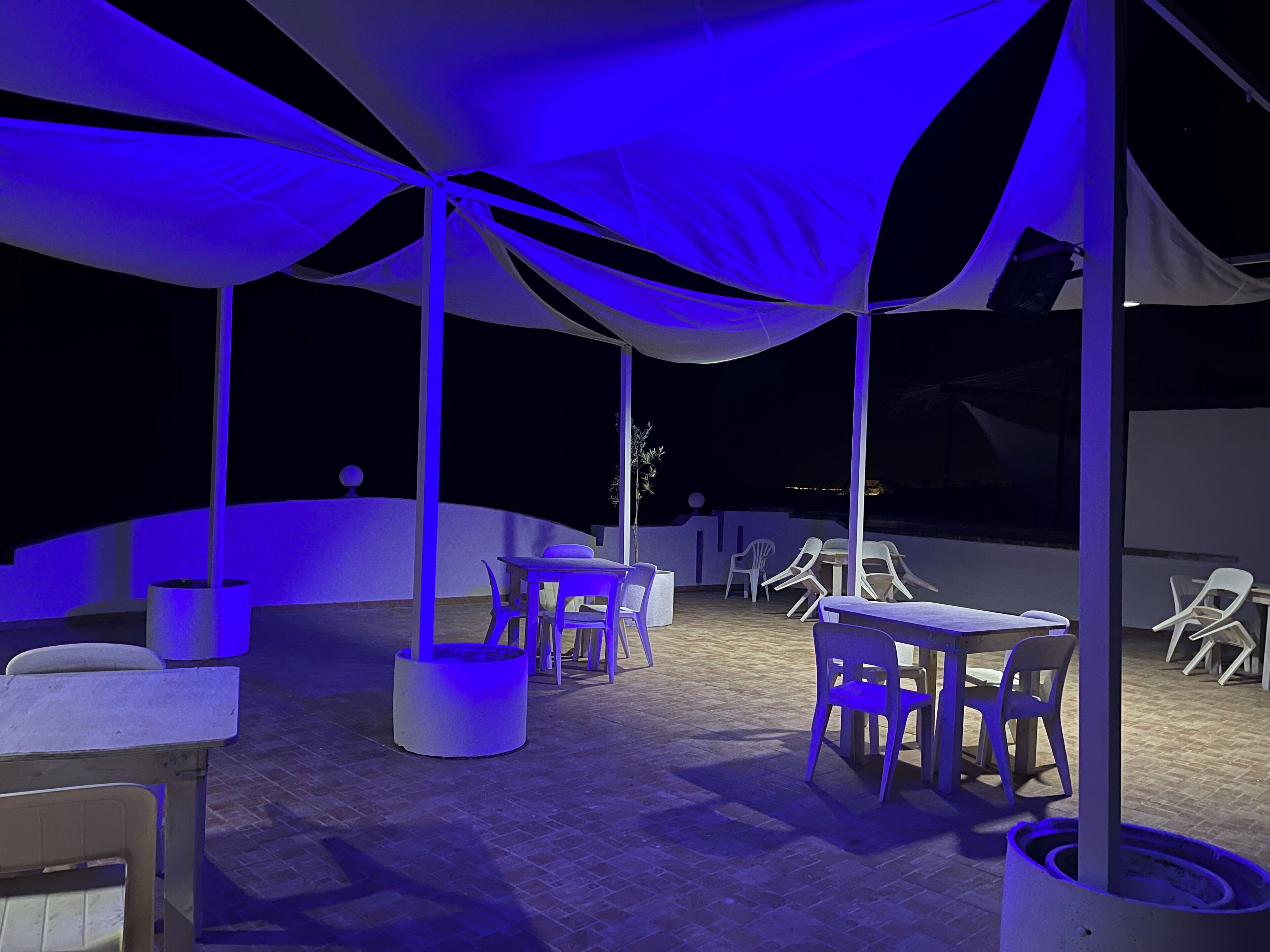 Un espacio de patio al aire libre con mesas y sillas bajo una gran carpa, con piso de ladrillo y mobiliario blanco.