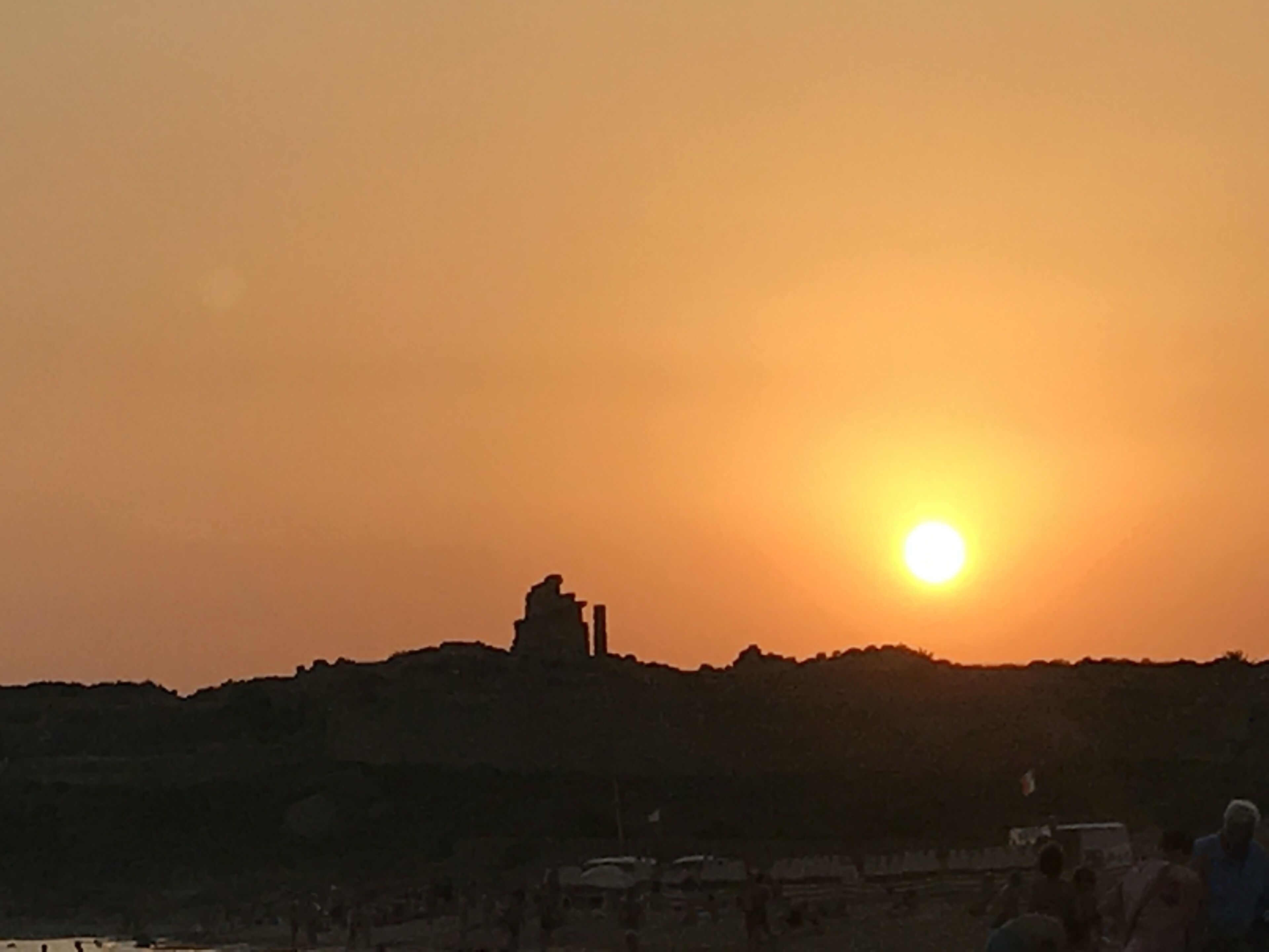 A sunset over a hilly landscape with a silhouette of a building and people in the foreground.