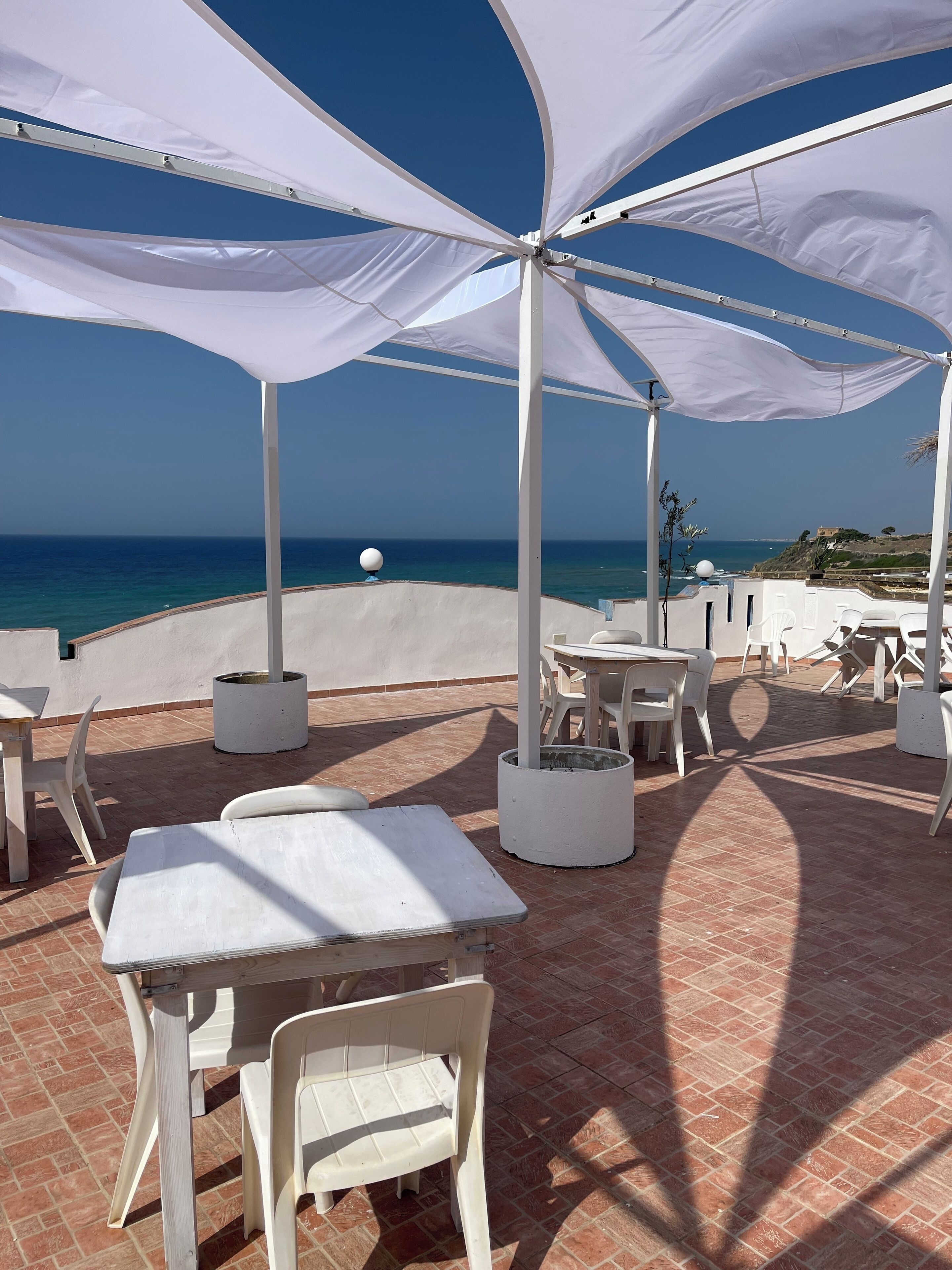 Un espace extérieur aménagé avec des tables et des chaises blanches, un auvent blanc et une vue sur la mer.