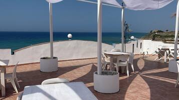 Un espace extérieur aménagé avec des tables et des chaises blanches, un auvent blanc et une vue sur la mer.