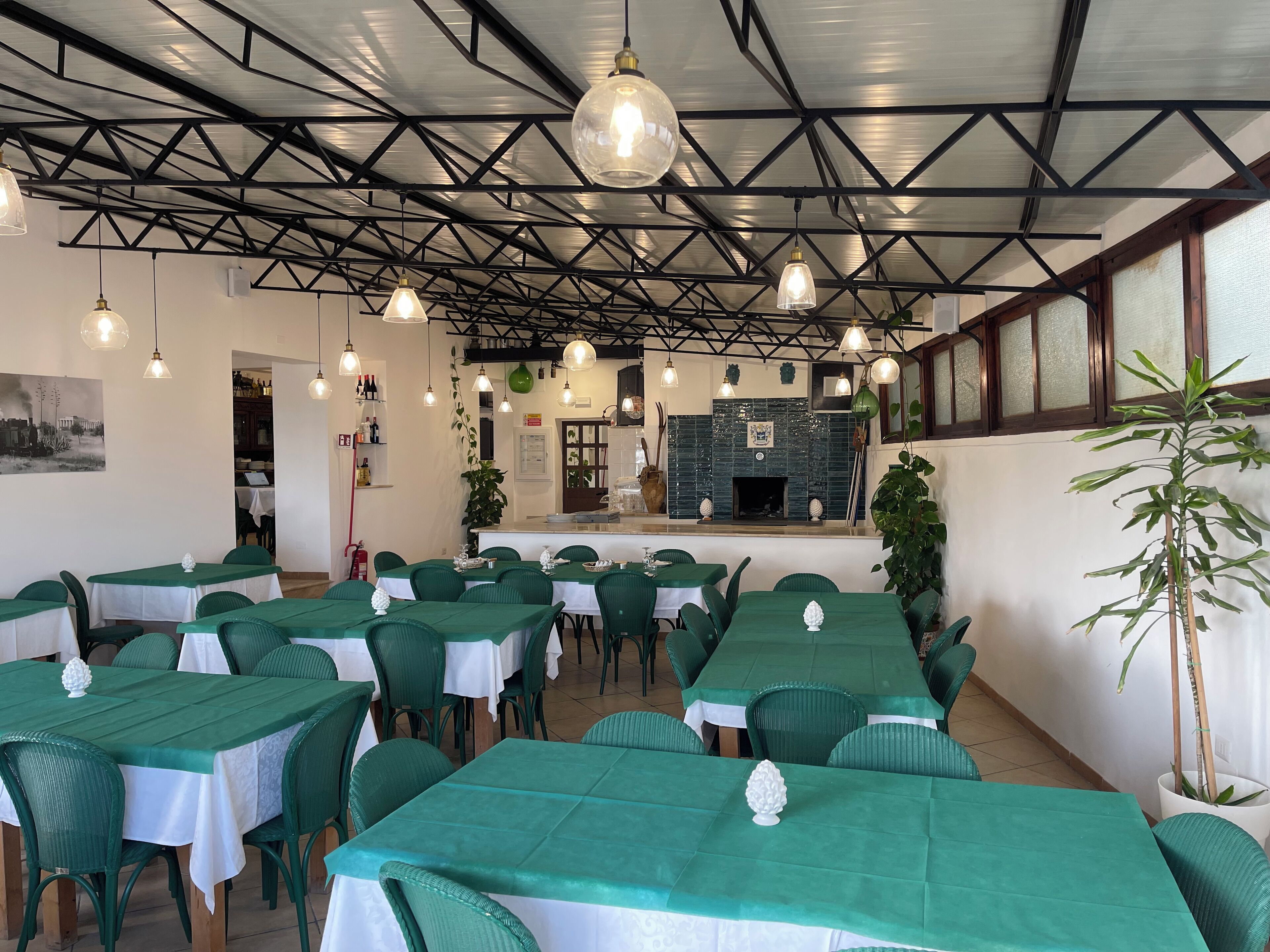 Un restaurant avec de nombreuses tables recouvertes de nappes vertes et de chemins de table blancs, agrémenté de luminaires suspendus et d’une cheminée en briques en arrière-plan.