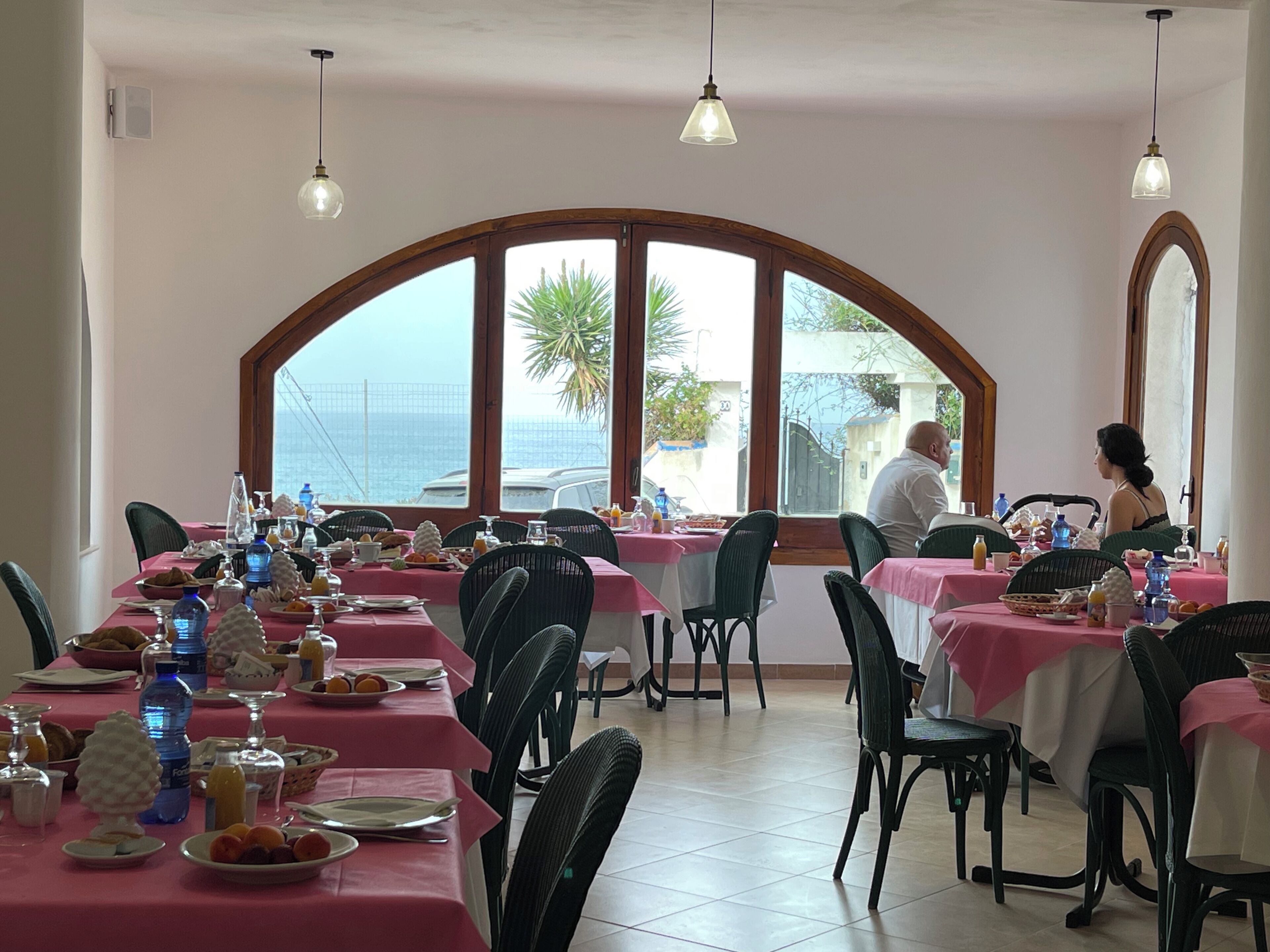 Comedor con mesas preparadas para el desayuno, ventanas amplias con vistas al mar y lámparas colgantes.