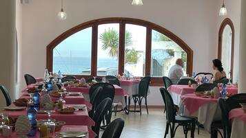 Comedor con mesas preparadas para el desayuno, ventanas amplias con vistas al mar y lámparas colgantes.