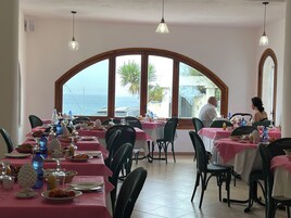 Comedor con mesas preparadas para el desayuno, ventanas amplias con vistas al mar y lámparas colgantes.