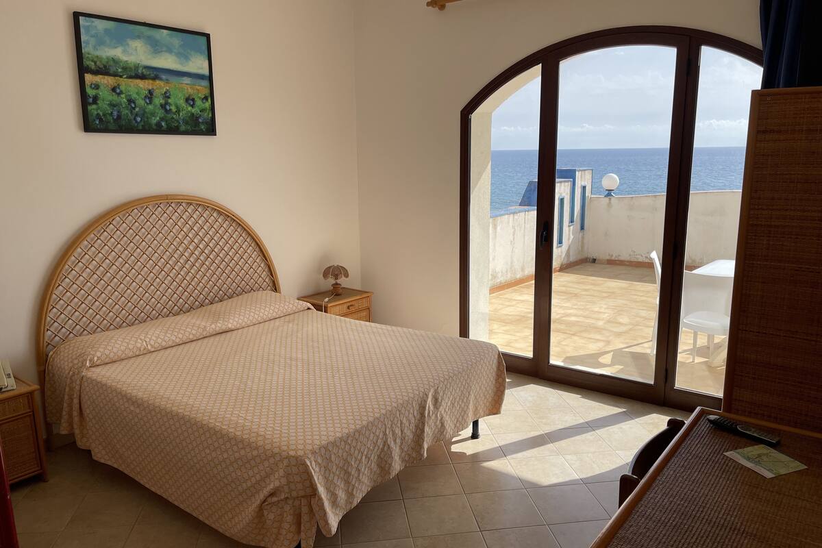 Une chambre à coucher avec un lit, une table de chevet, une lampe, un tableau et une vue sur la mer, visible à travers une porte vitrée.