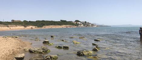 A beach with clear water, rocks, and buildings in the background.