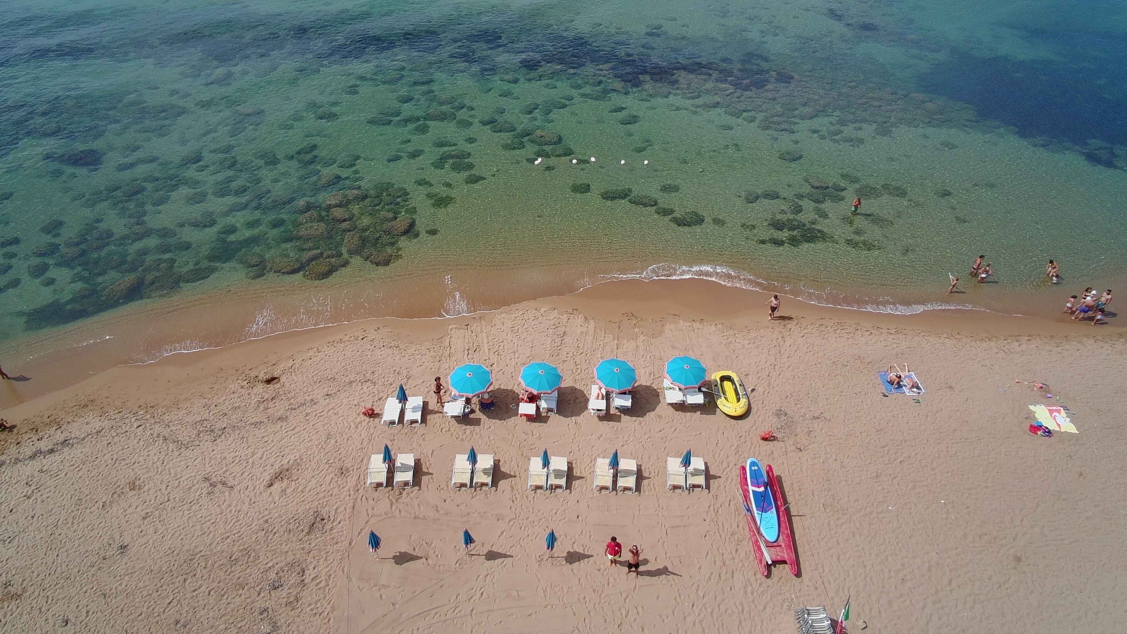 An aerial view of a beach with clear water, several beach umbrellas, lounge chairs, and a few people.