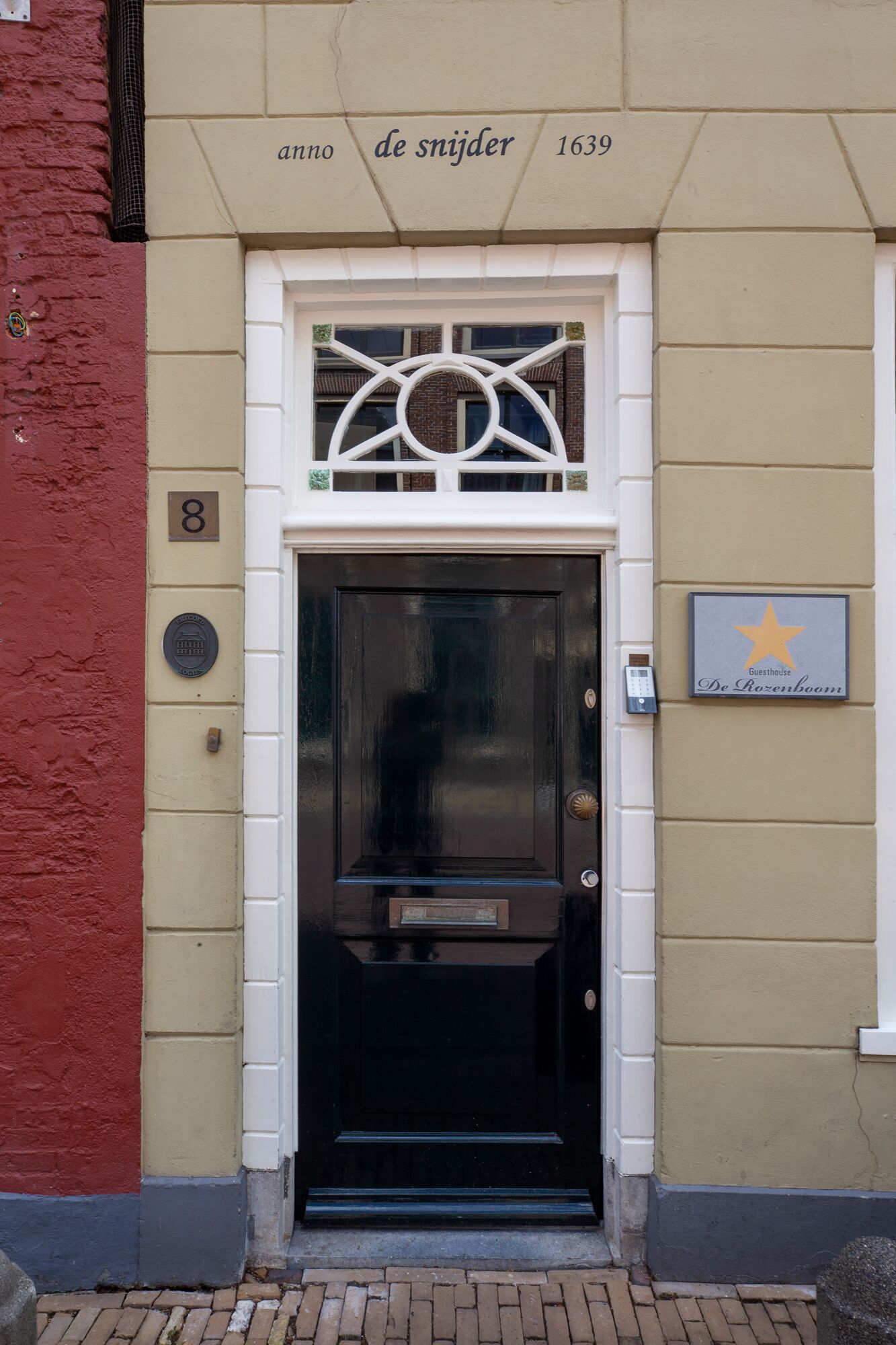 Una puerta negra con un marco decorativo blanco, una placa con el año 1639 y un cartel que indica 'Guesthouse De Rozenboom'.