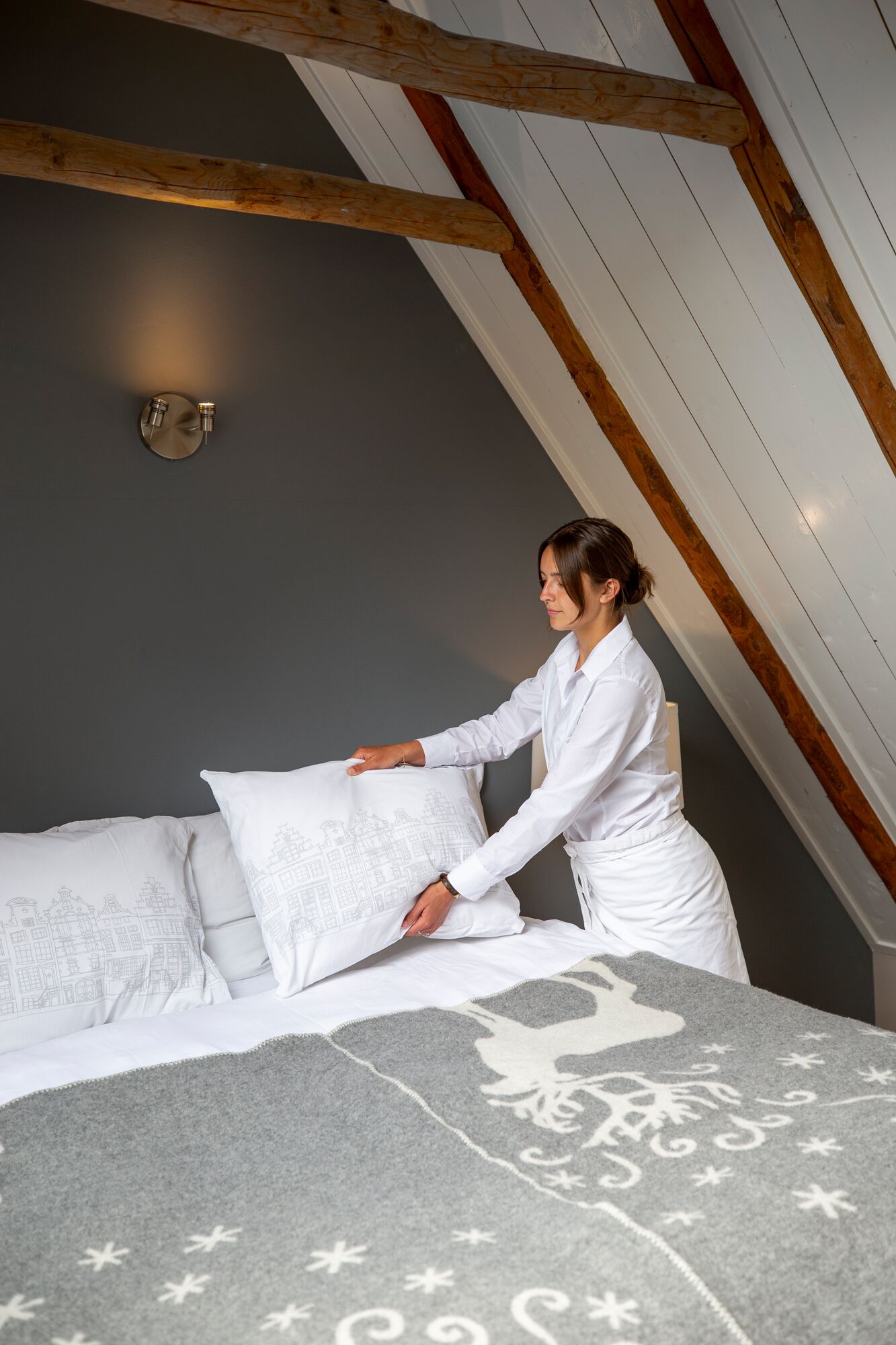Eine Person richtet ein Bett in einem Zimmer mit schrägem Dach und freiliegenden Holzbalken ein.
