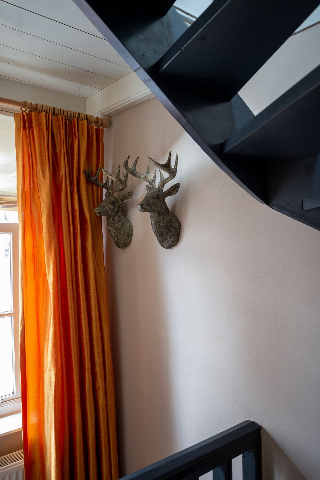 Un escalier avec une rampe noire, une décoration murale représentant une tête de cerf et un rideau orange.