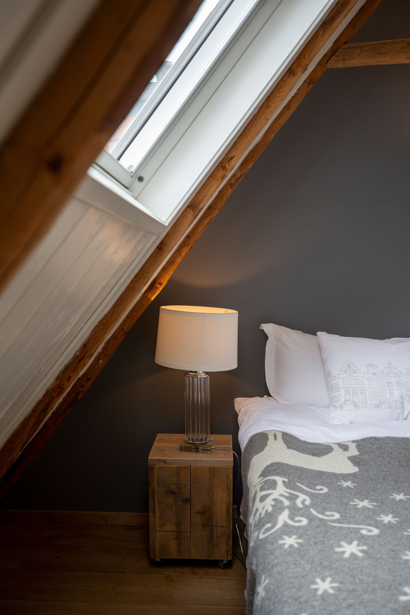 Un dormitorio con tragaluz, una lámpara de noche, una cama con ropa de cama blanca y una manta decorativa.