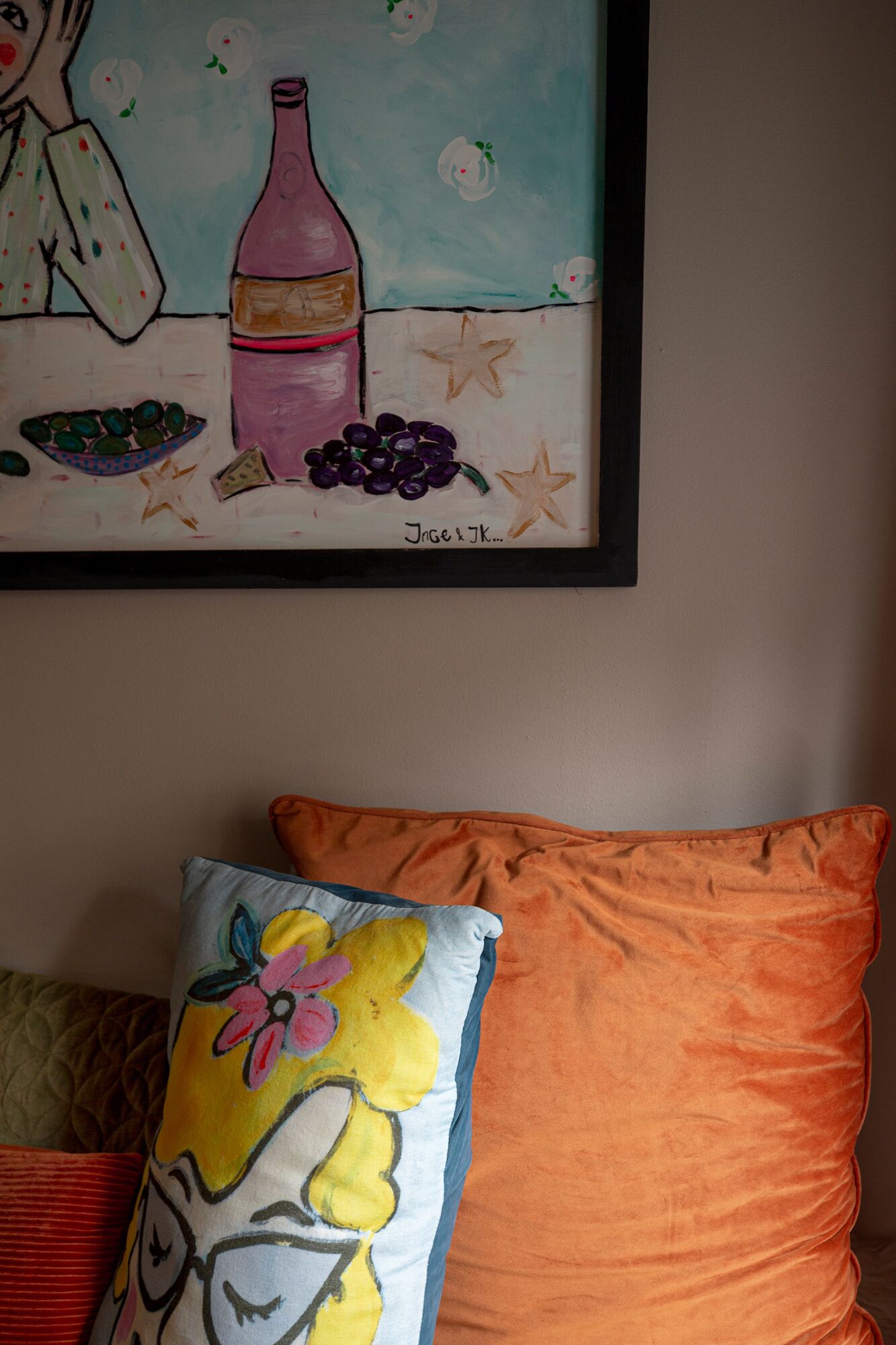 Un tableau encadré représentant une femme tenant une bouteille, un bol d’olives et des raisins, le tout accroché à un mur.